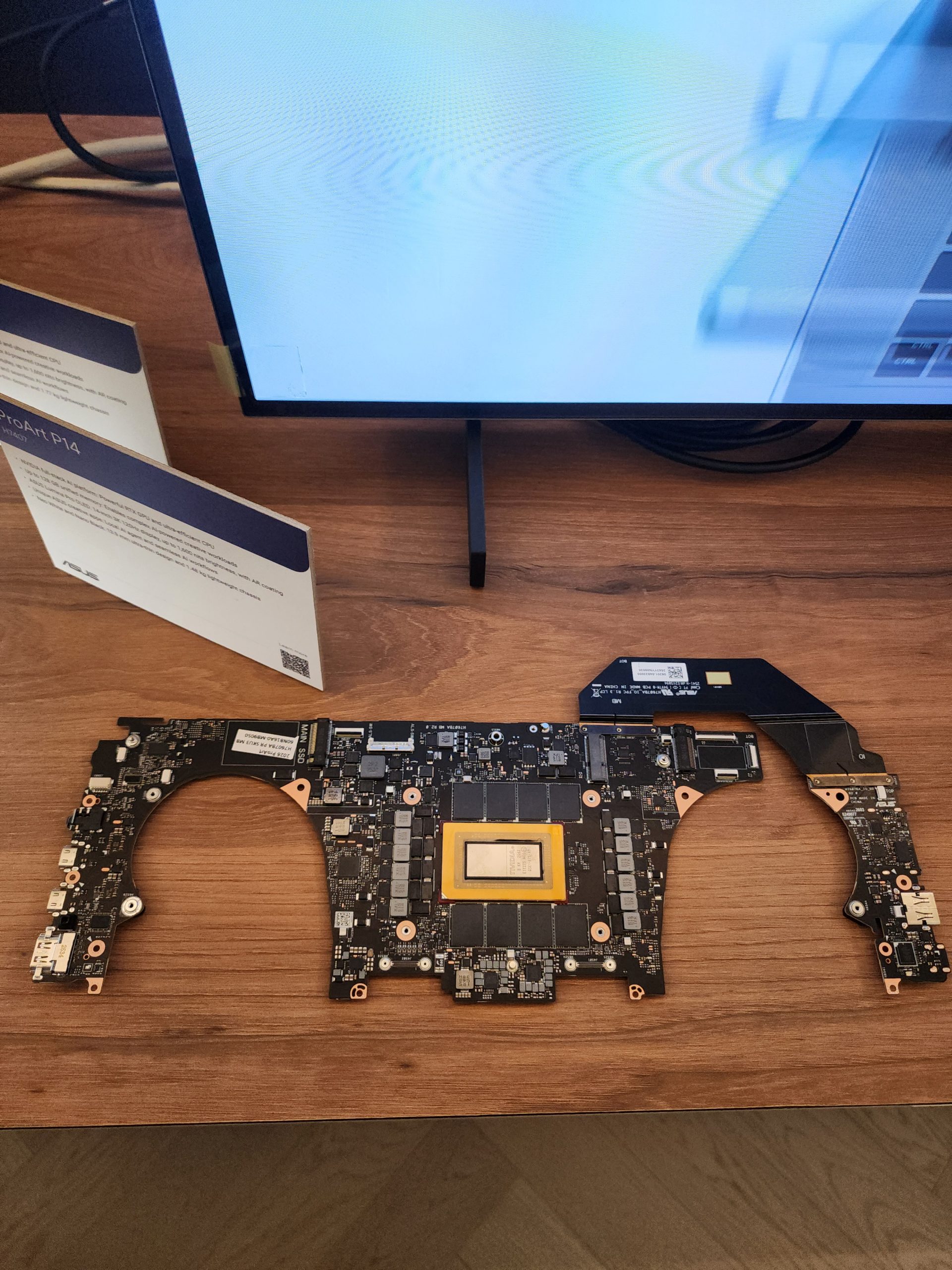 An ASUS motherboard labeled 'ProArt P14' is displayed on a wooden surface, with a monitor showing a blue and white abstract pattern above it.