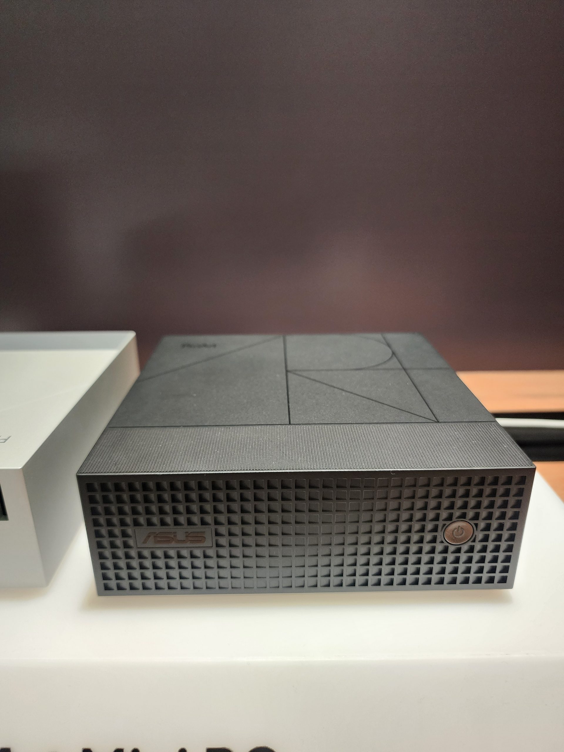 A black ASUS mini PC with a textured front panel and power button is displayed on a white surface.