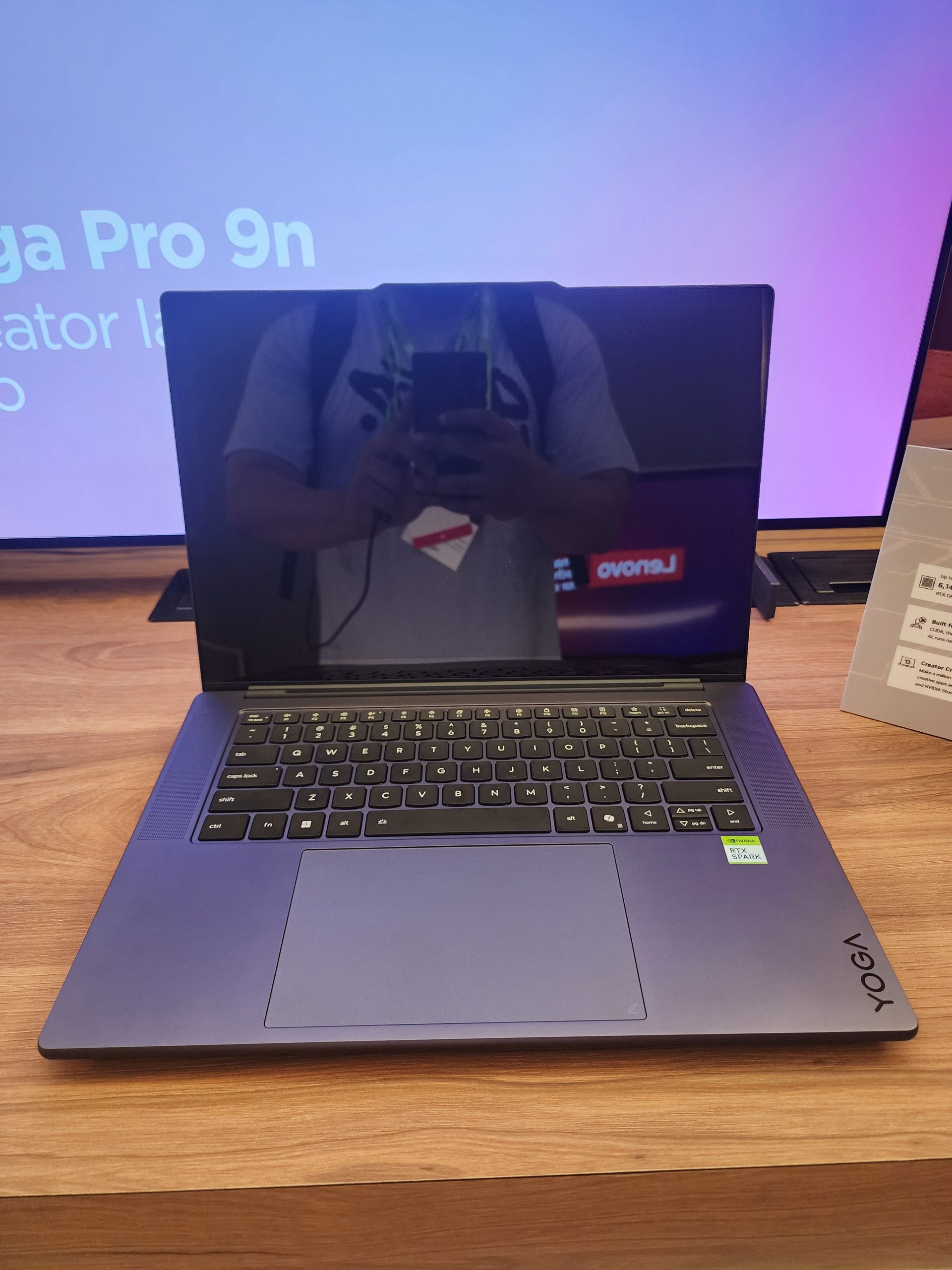 A Lenovo Yoga Pro 9n laptop on display with its screen off, reflecting a person taking a photo and showing the 'RTX Studio' sticker.