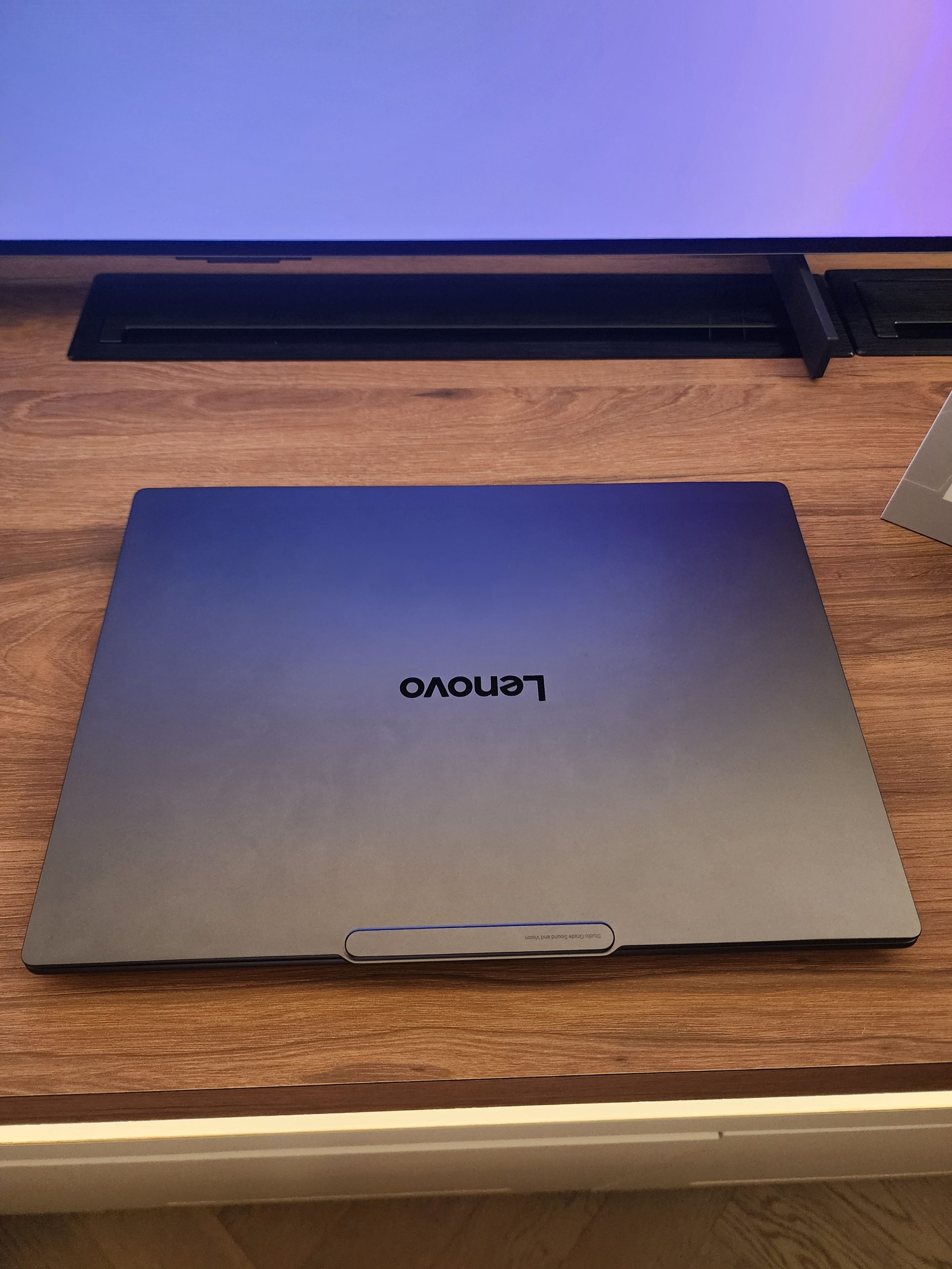 A Lenovo laptop is closed on a wooden desk in front of a television screen.