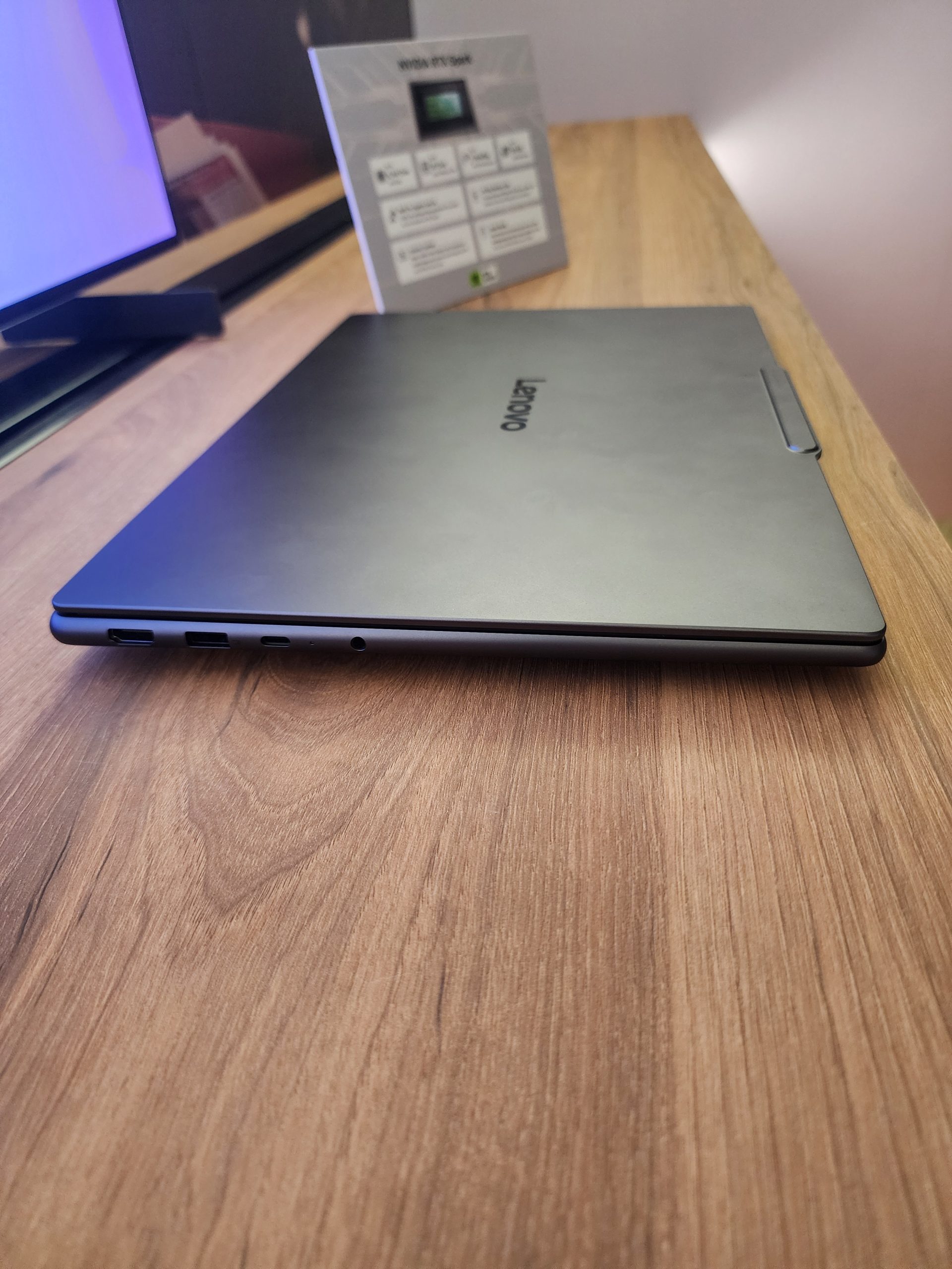 A closed Lenovo laptop is on a wooden table next to a stand displaying 'NVIDIA RTX 5080' with specifications.