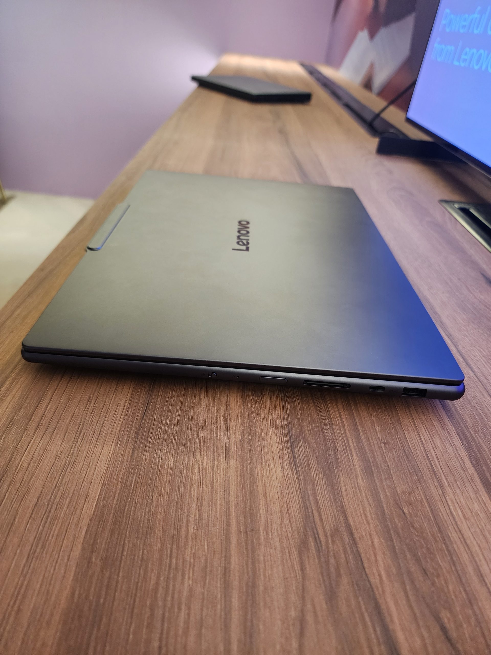 A closed Lenovo laptop is placed on a wooden surface next to another device and a monitor displaying 'Powerful Innovation from Lenovo'.