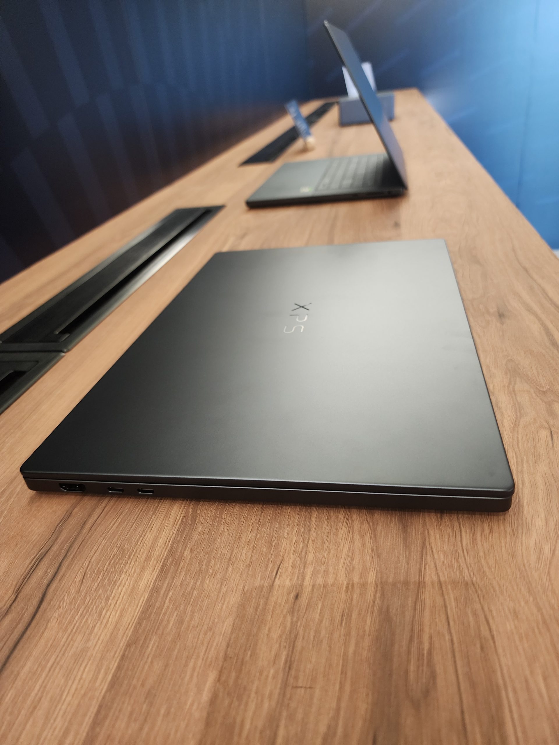 A Dell XPS laptop with a closed lid is displayed on a wooden table, with other laptops in the background.