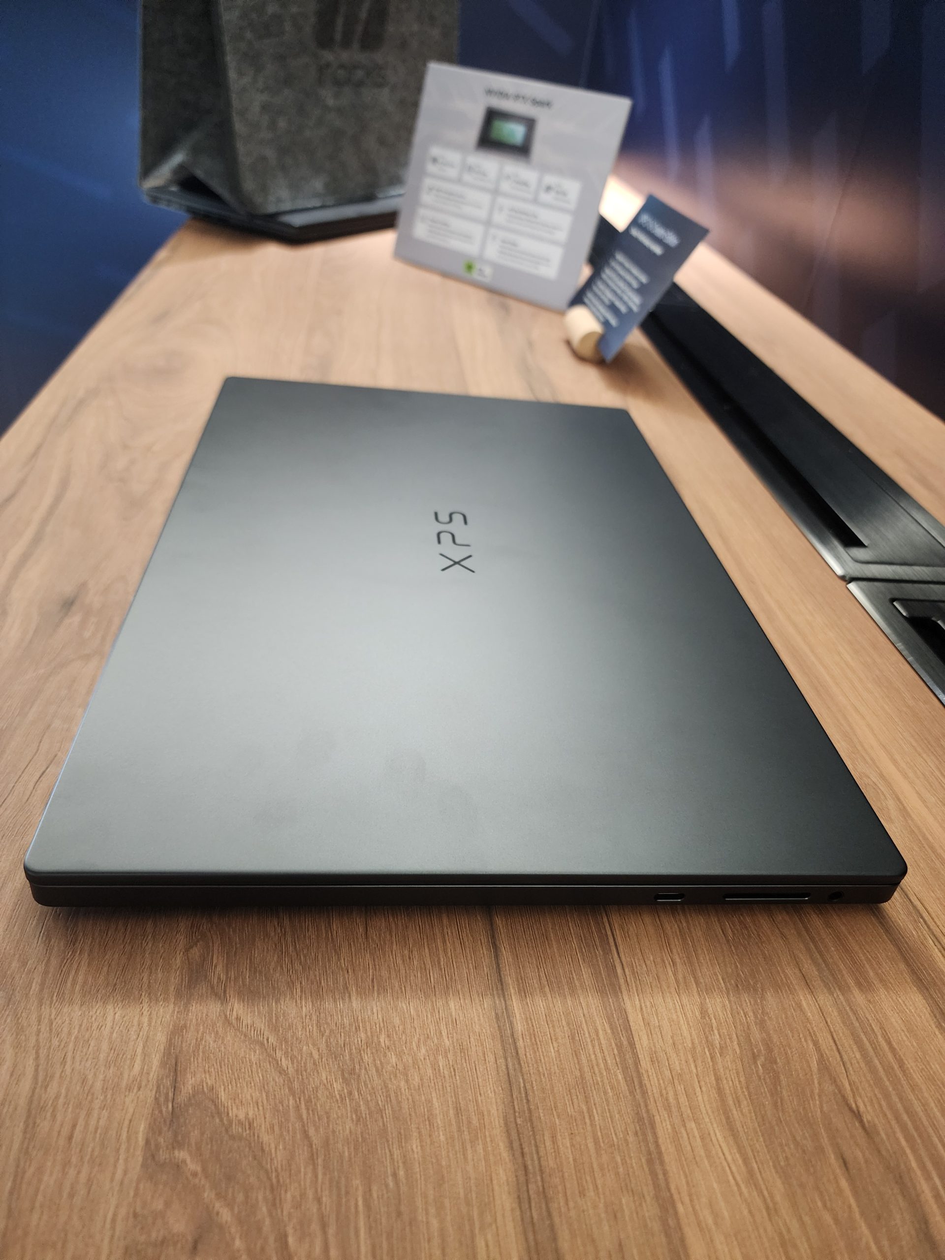 A Dell XPS laptop closed on a wooden table with a product specification card in the background.