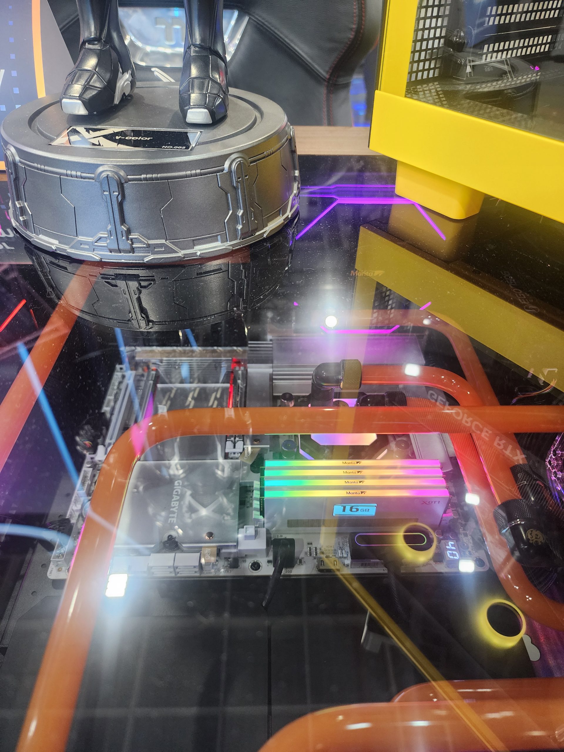 A GIGABYTE motherboard setup with colorful RGB lighting, featuring MAIYU JFL DDR RAM labeled '16GB,' surrounded by custom orange liquid cooling tubes and a futuristic showpiece on top.