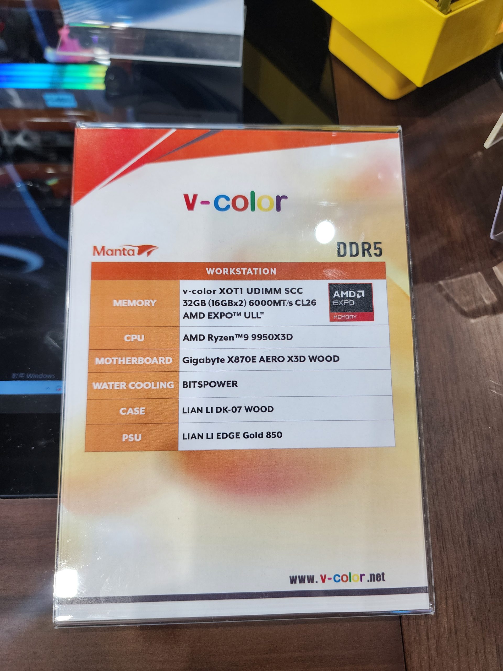 A product information sign for a v-color workstation lists components including 'v-color XOT1 UDIMM SCC 32GB (16GBx2) 6000MT/s CL26 AMD EXPO™ ULL,' AMD Ryzen™ 9 9950X3D CPU, Gigabyte X870E AERO X3D WOOD motherboard, and LIAN LI DK-07 WOOD case.