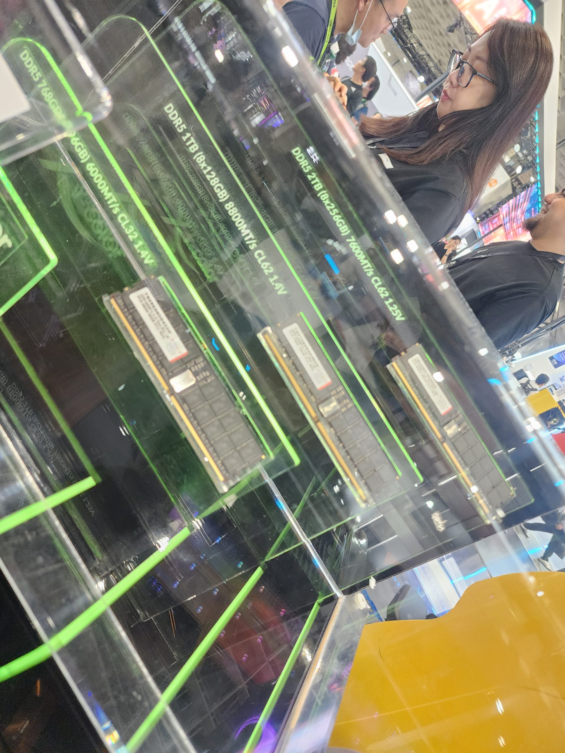 A display case showcasing DDR5 RAM modules with specifications such as 'DDR5-1TB (8x128GB) 8800MT/s CL62-AV' at a tech event with people in the background.