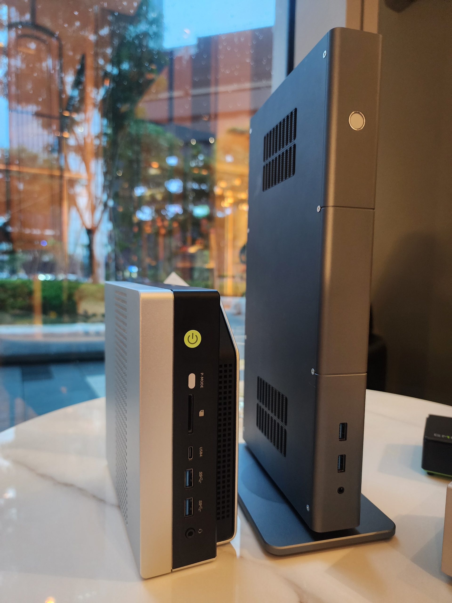 Two compact, unbranded mini PC cases side by side on a table, showcasing various front and side ports, with a windowed urban background.