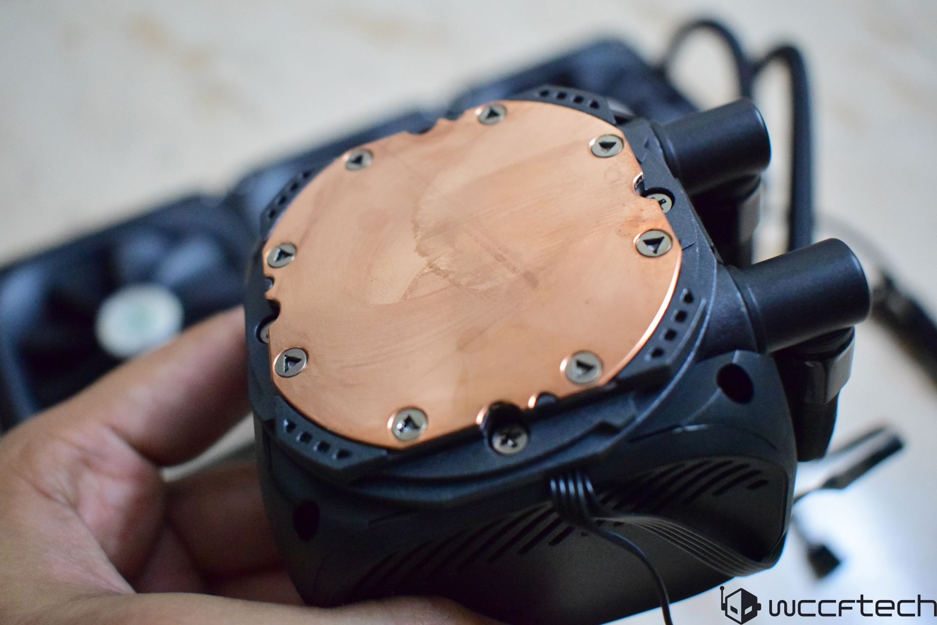 A hand holds an unbranded liquid CPU cooler, showing its copper base plate with screws, against a blurred background featuring a radiator and fan.