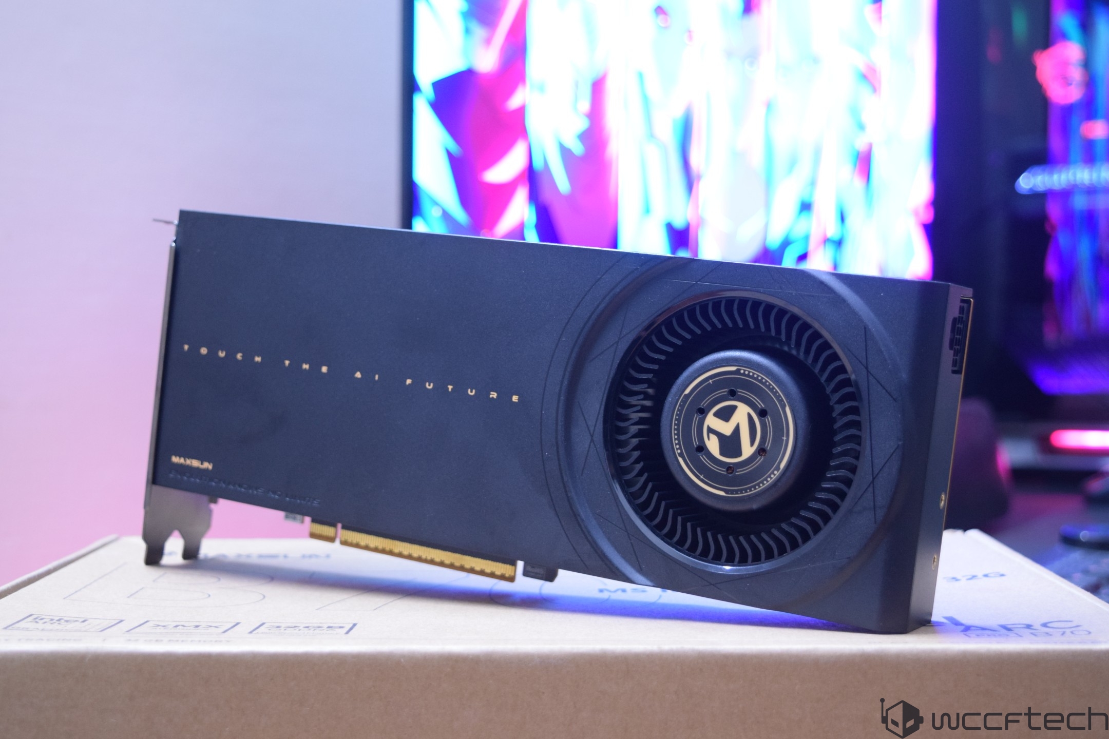 A Maxsun graphics card with the text 'TOUCH THE AI FUTURE' printed on it is displayed on top of a cardboard box, with a colorful monitor screen in the background.