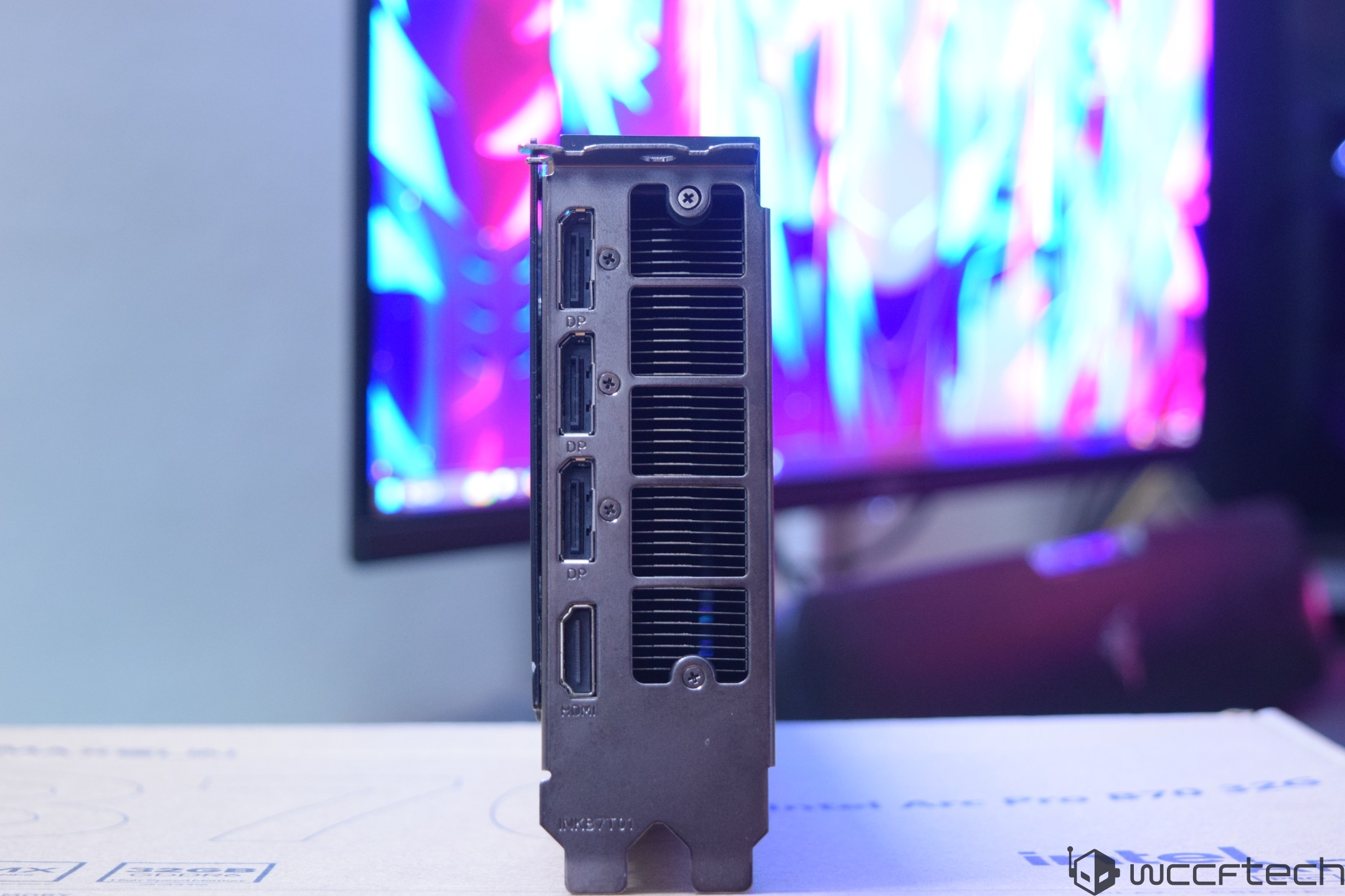 The image shows the back panel of a graphics card with four DisplayPort (DP) connections and one HDMI port, positioned on a desk in front of a blurred monitor displaying vibrant colors.