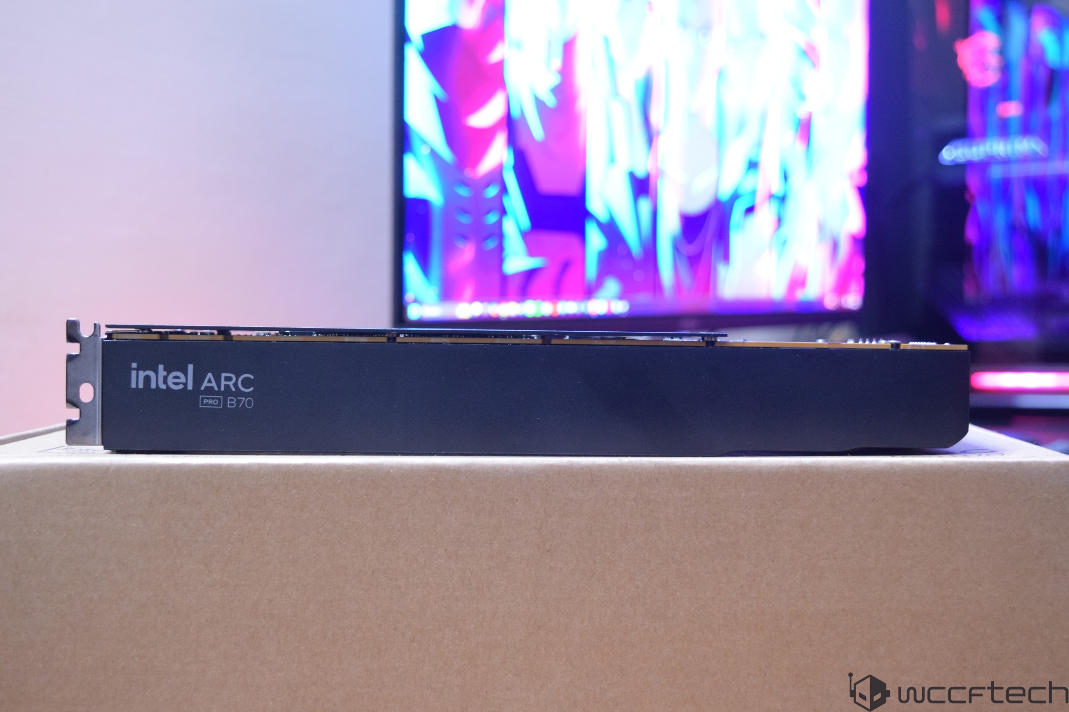 A close-up of an Intel ARC Pro B70 graphics card resting on a cardboard box with colorful screens in the background.
