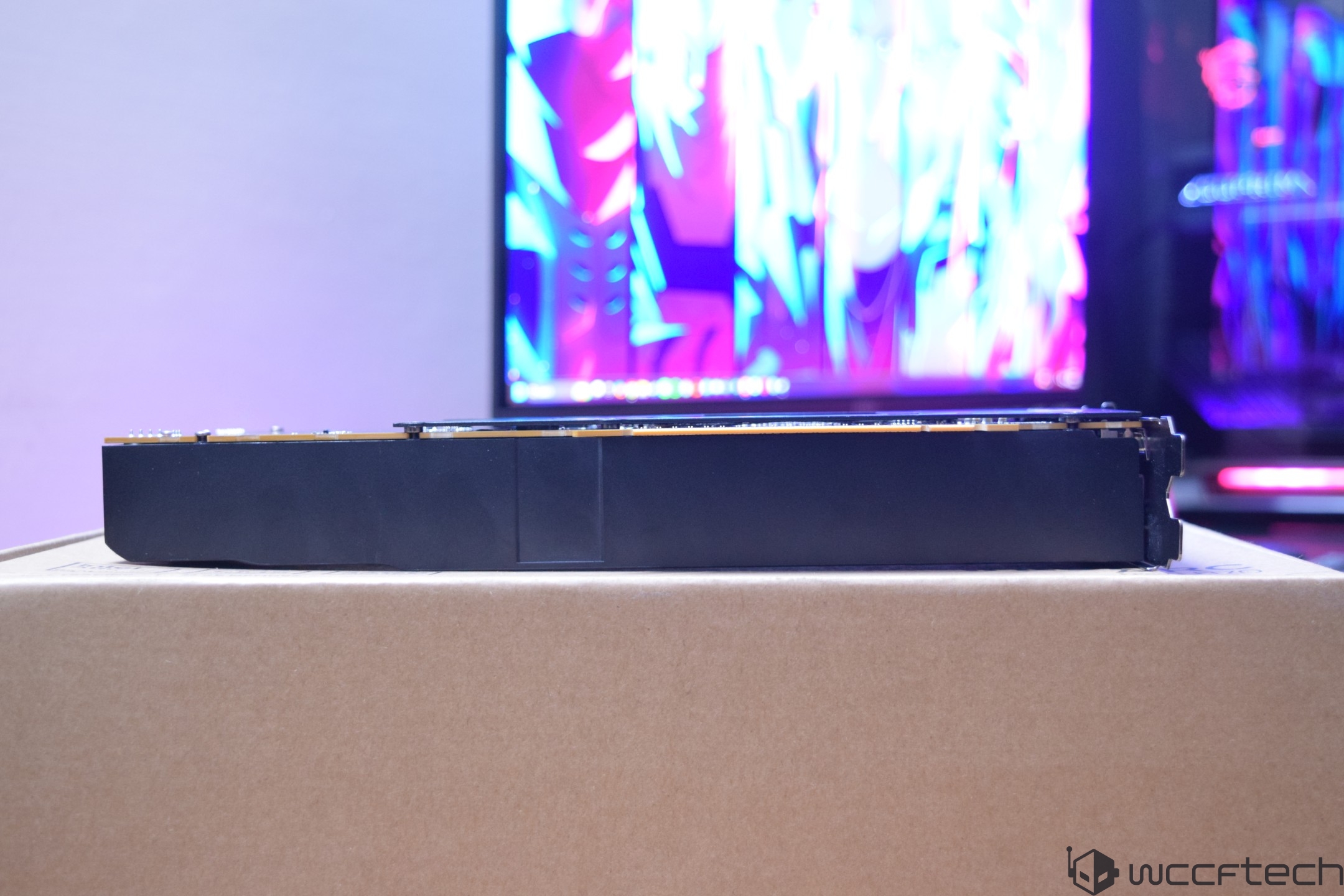 A horizontal view of an unbranded black graphics card resting on a cardboard box, with a colorful abstract image displayed on a screen in the background.