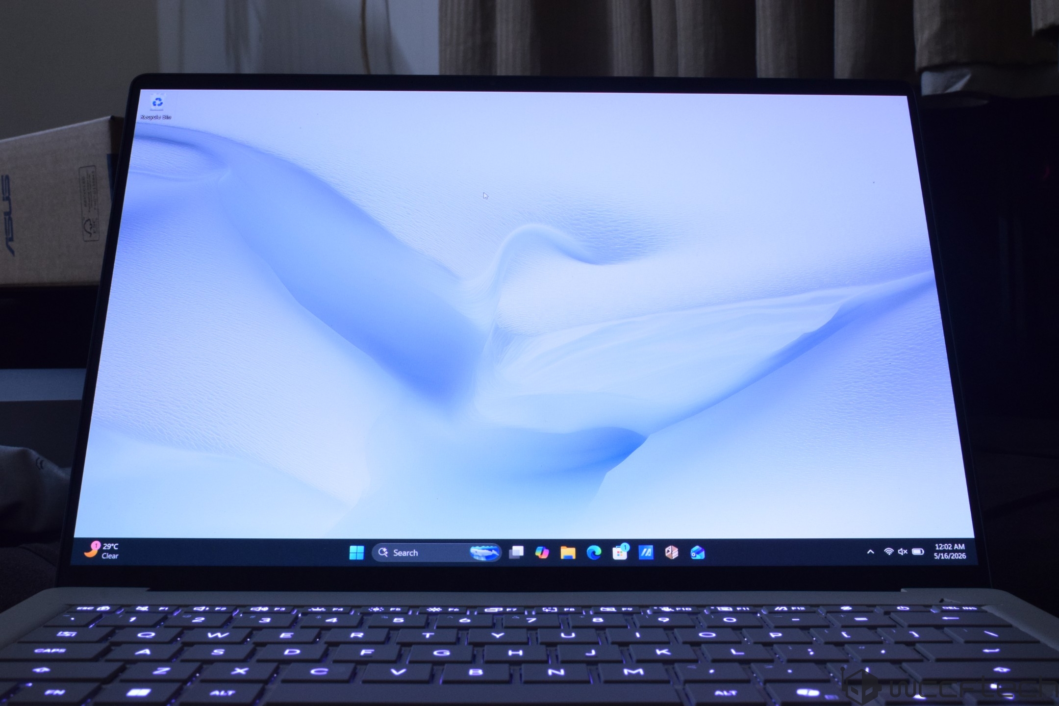 An open ASUS laptop displays a desktop with a 'Recycling Bin' icon, Windows taskbar, and the time '12:02 AM' on '5/16/2026'.