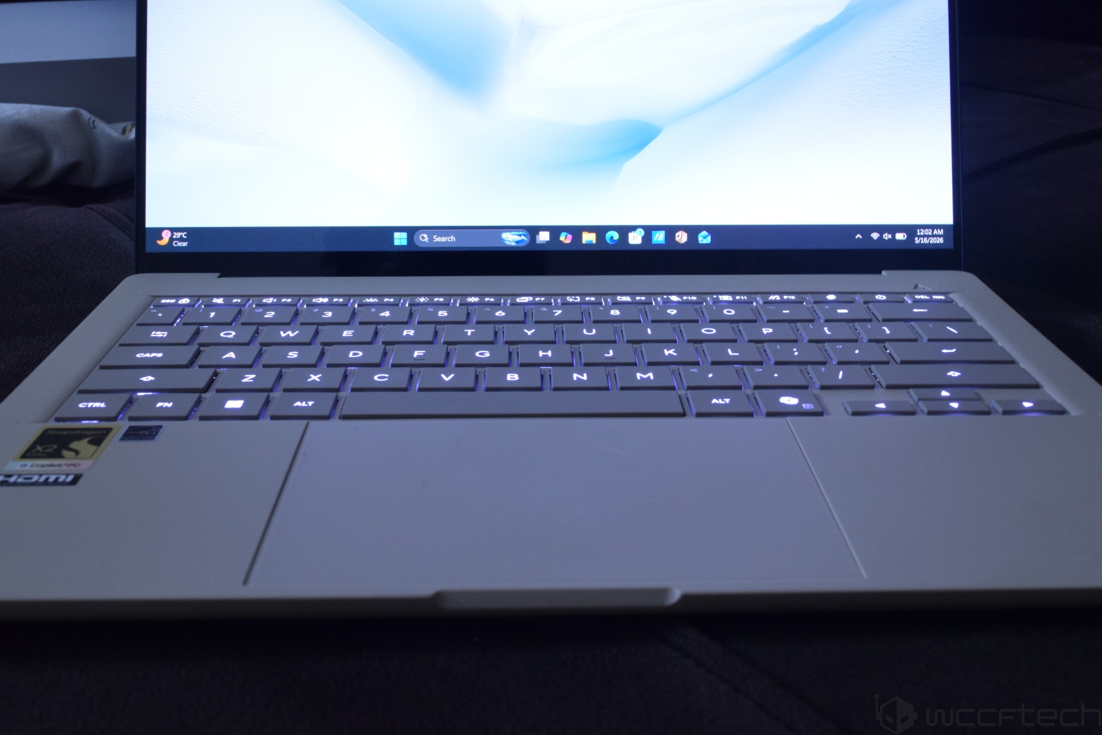 A white laptop with backlit keys and a visible 'Qualcomm Snapdragon' sticker on the touchpad displaying a Windows desktop interface with icons and a '37°C Clear' weather notification.