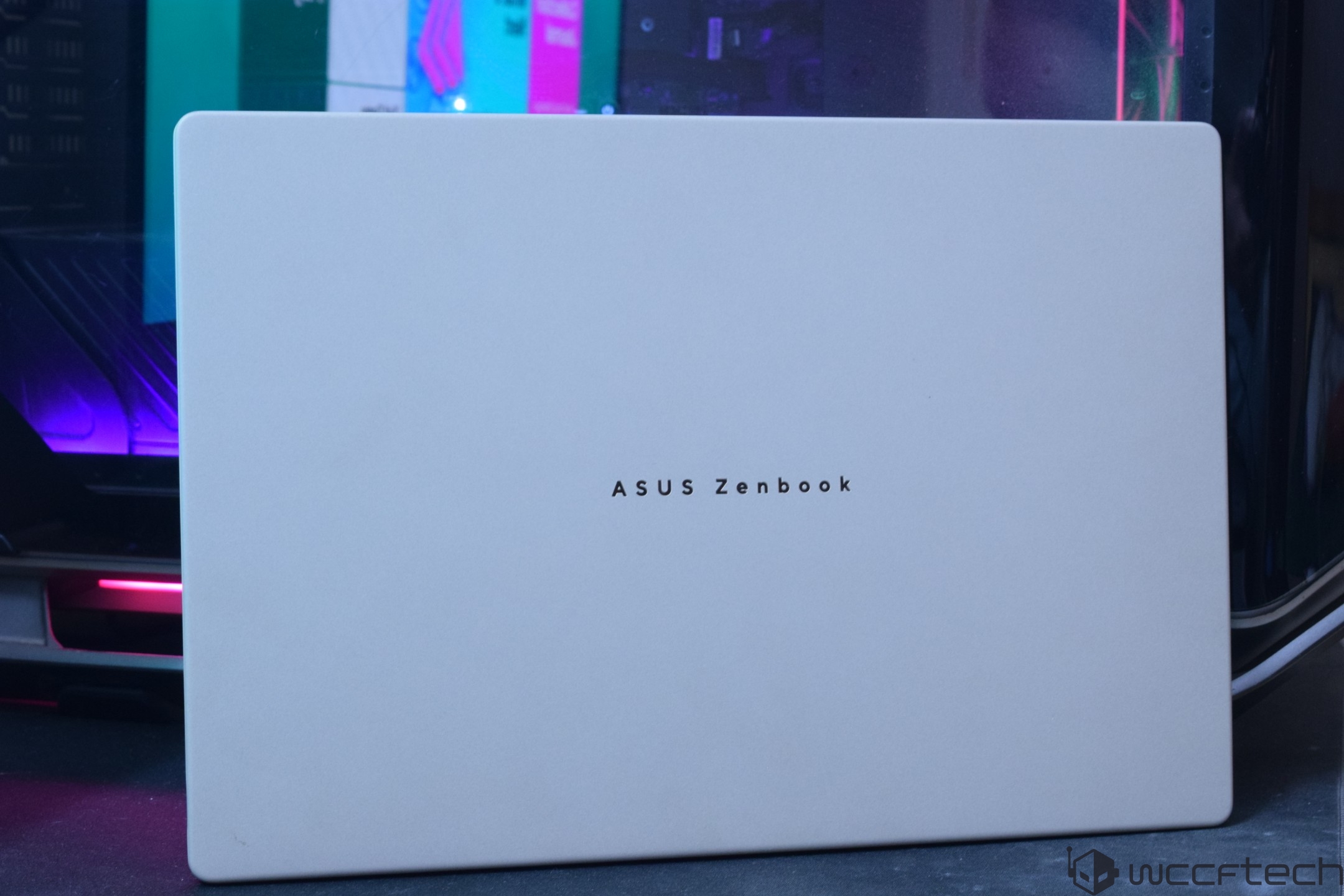 The image shows an ASUS Zenbook laptop with its lid closed, placed in front of a desktop setup with RGB lighting.
