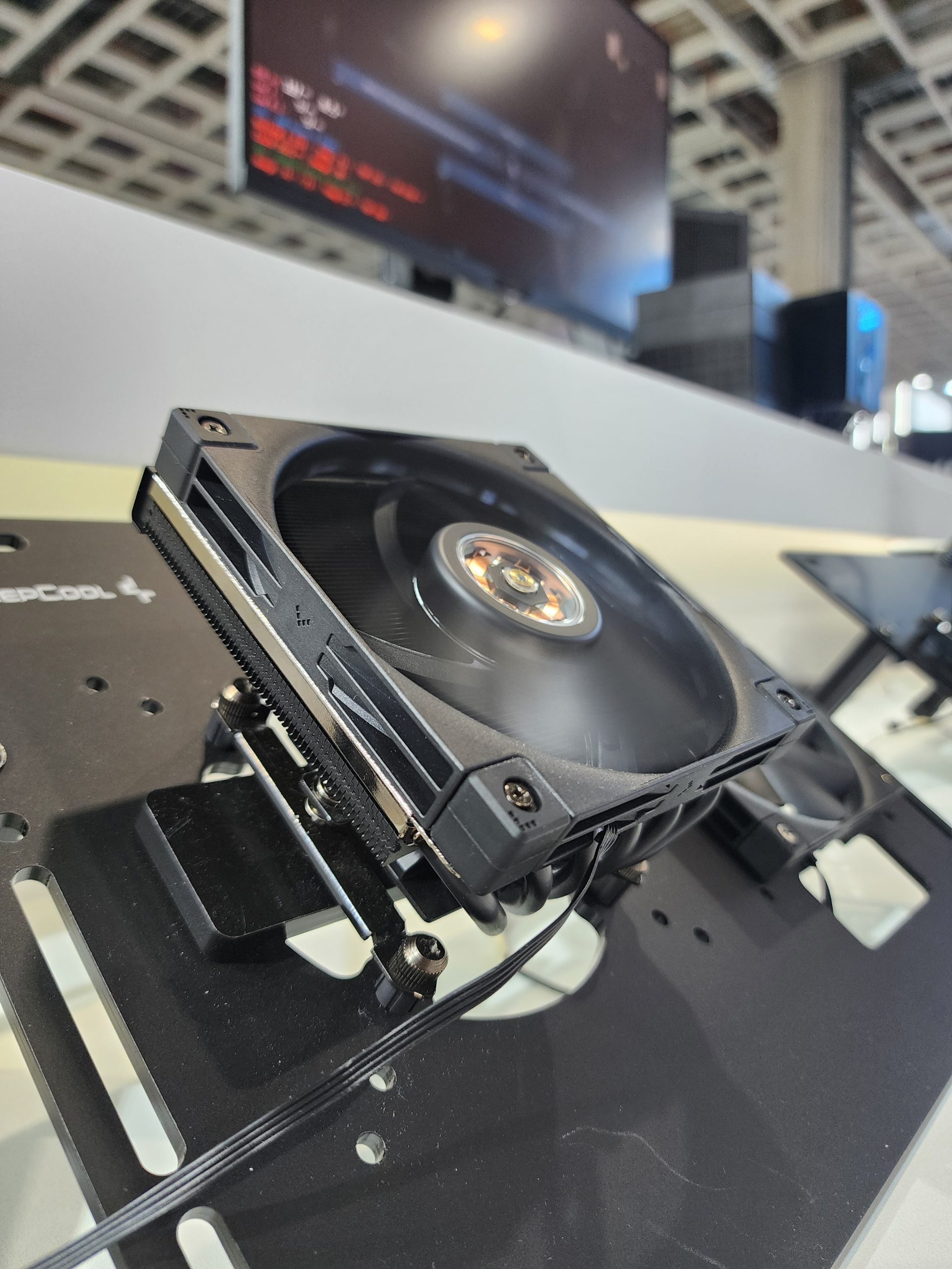 A DeepCool fan is mounted on a test bench with a blurred screen displaying lines of code in the background.