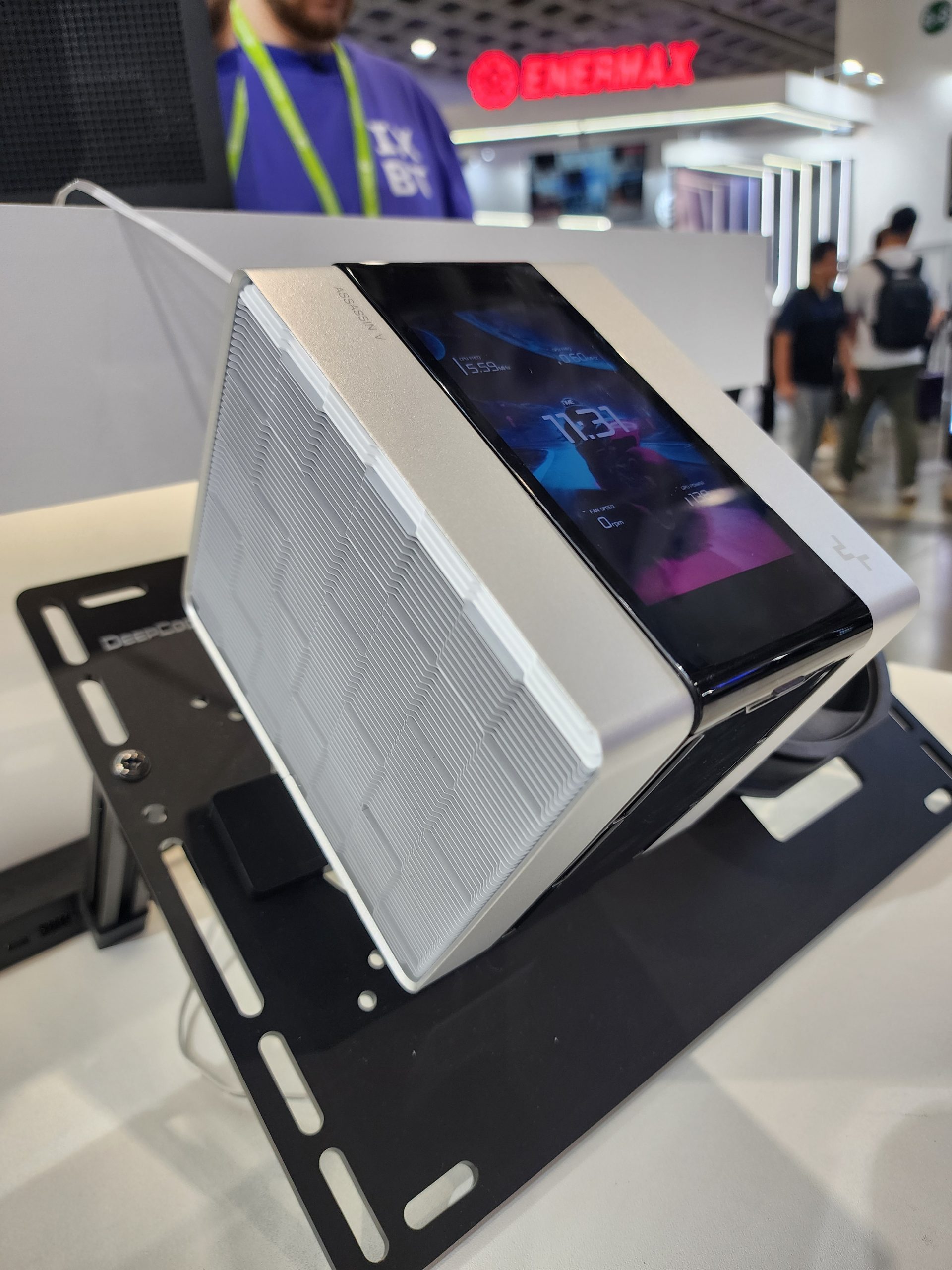 Unbranded triangular PC case with an integrated screen displaying '13:19' and '1555 RPM', exhibited at a tech event with an Enermax sign visible in the background.