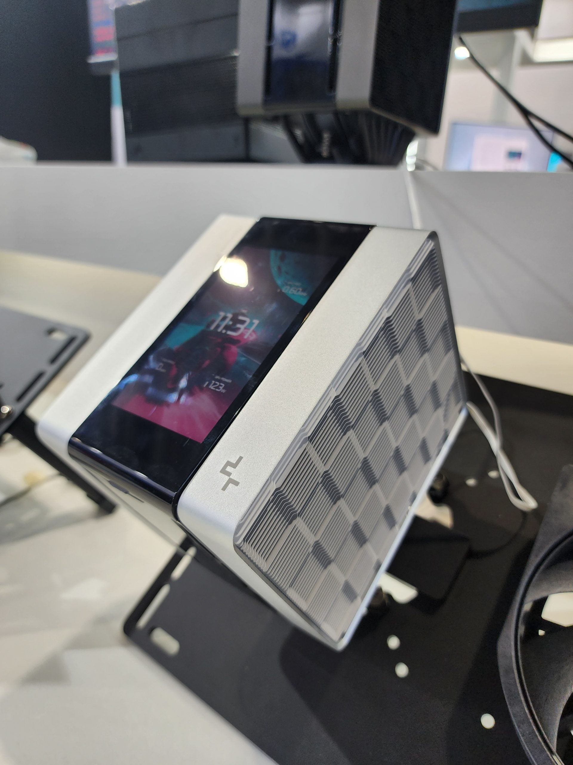 A compact PC with a built-in screen displaying '11:37' is housed in a sleek, silver chassis with a geometric pattern and a logo on the corner.