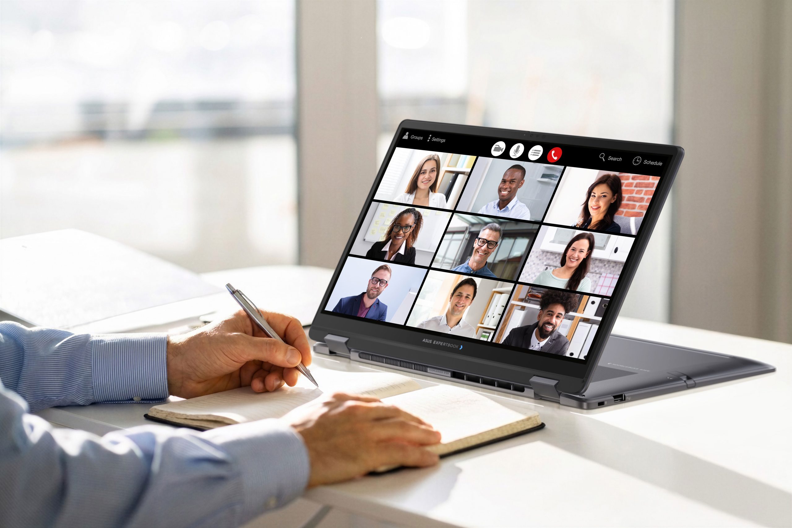 A person takes notes while participating in a video call on an ASUS ExpertBook laptop showing nine participants.