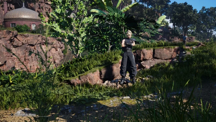 A character from 'Final Fantasy VII Rebirth' stands in a lush, forested area with rock formations and a round building in the background.