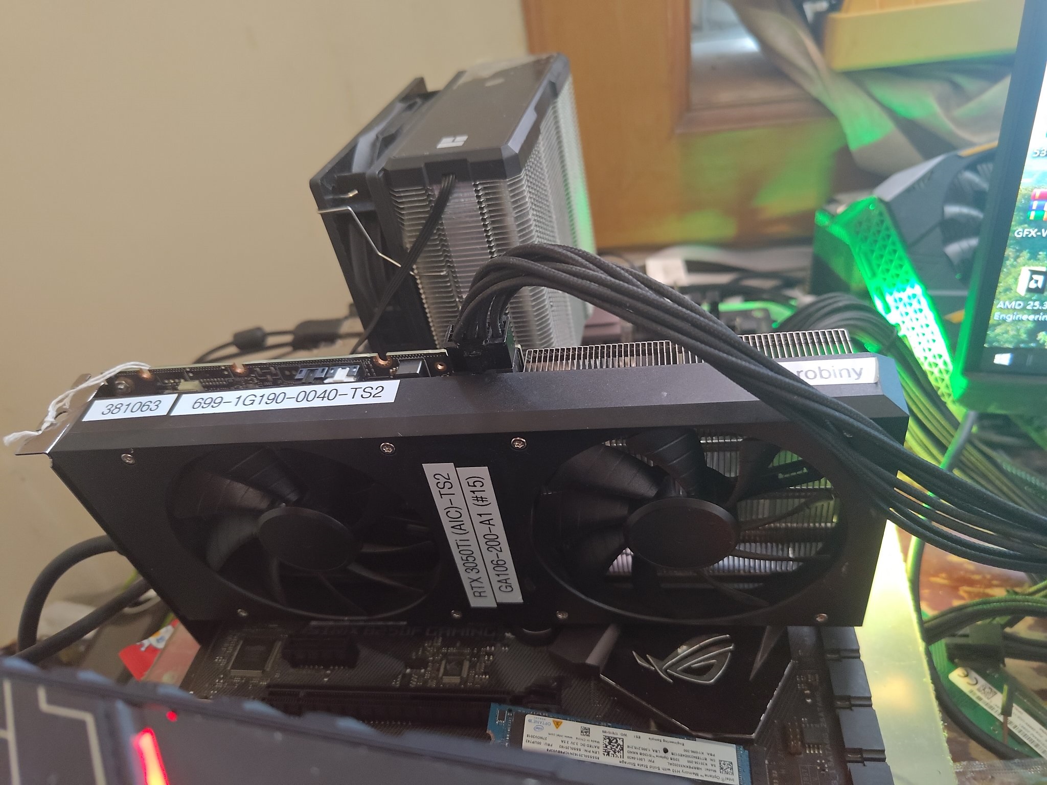 A graphics card labeled RTX 3050 TI (AIC) - TS2 is installed in an Asus ROG motherboard with a visible large heatsink cooler in the background.