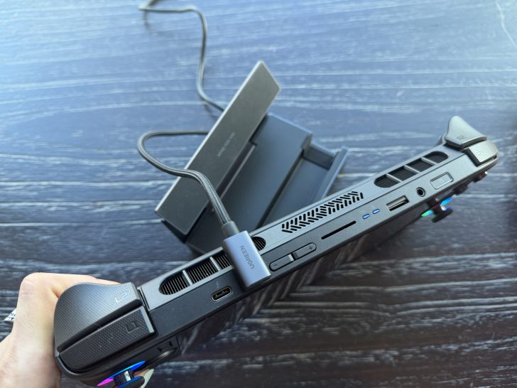 A handheld gaming device plugged into a UGREEN cable, showing ports and air vents against a dock on a wooden surface.