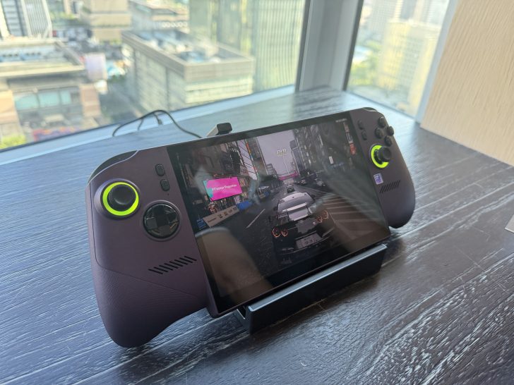 A handheld gaming device displays a racing game with on-screen text '#FasterTogether' by a window overlooking a cityscape.