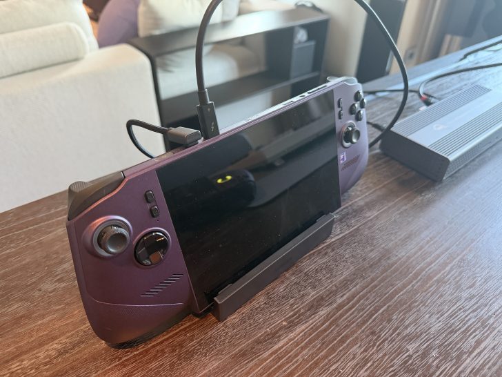 An unbranded handheld gaming console with a purple finish is docked on a table, connected via a USB-C cable.