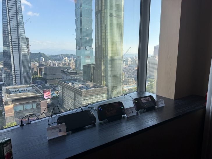 Three handheld gaming devices, including two labeled OneXplayer 2 and Acer Predator, are displayed on a window ledge overlooking a cityscape with the Taipei 101 building.