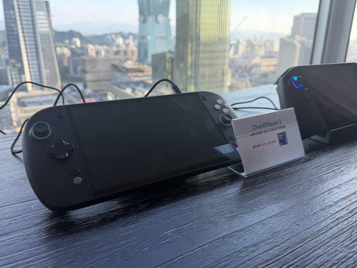 A OneXPlayer 3 handheld gaming device is displayed on a table with a sign reading 'OneXPlayer 3 with Intel Arc GXEXTREME' next to a window overlooking a city skyline.