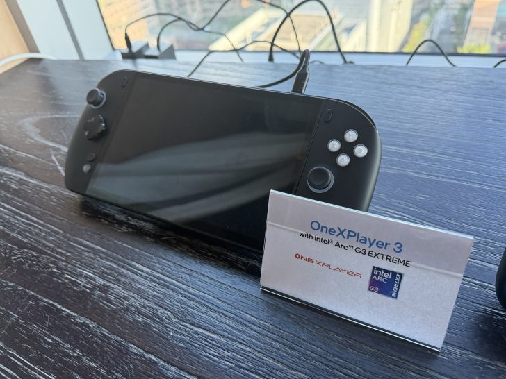 The OneXPlayer 3, a handheld gaming device, is displayed on a table alongside a sign indicating it features the Intel Arc G3 Extreme.