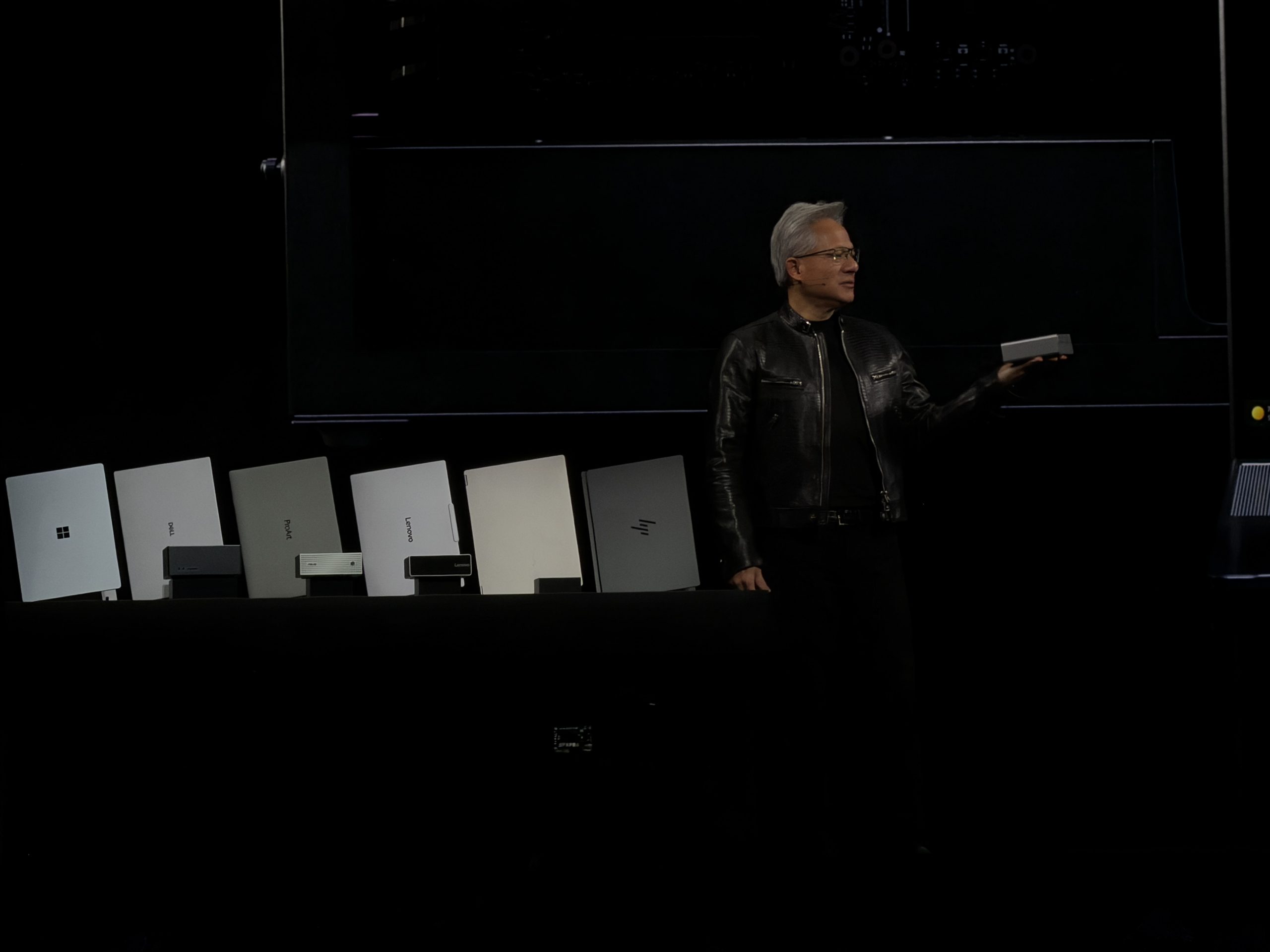 A person in a leather jacket holds a small electronic device while standing next to a row of various brand-name laptops, including Microsoft, ASUS, Dell, and Lenovo, on a dark stage.