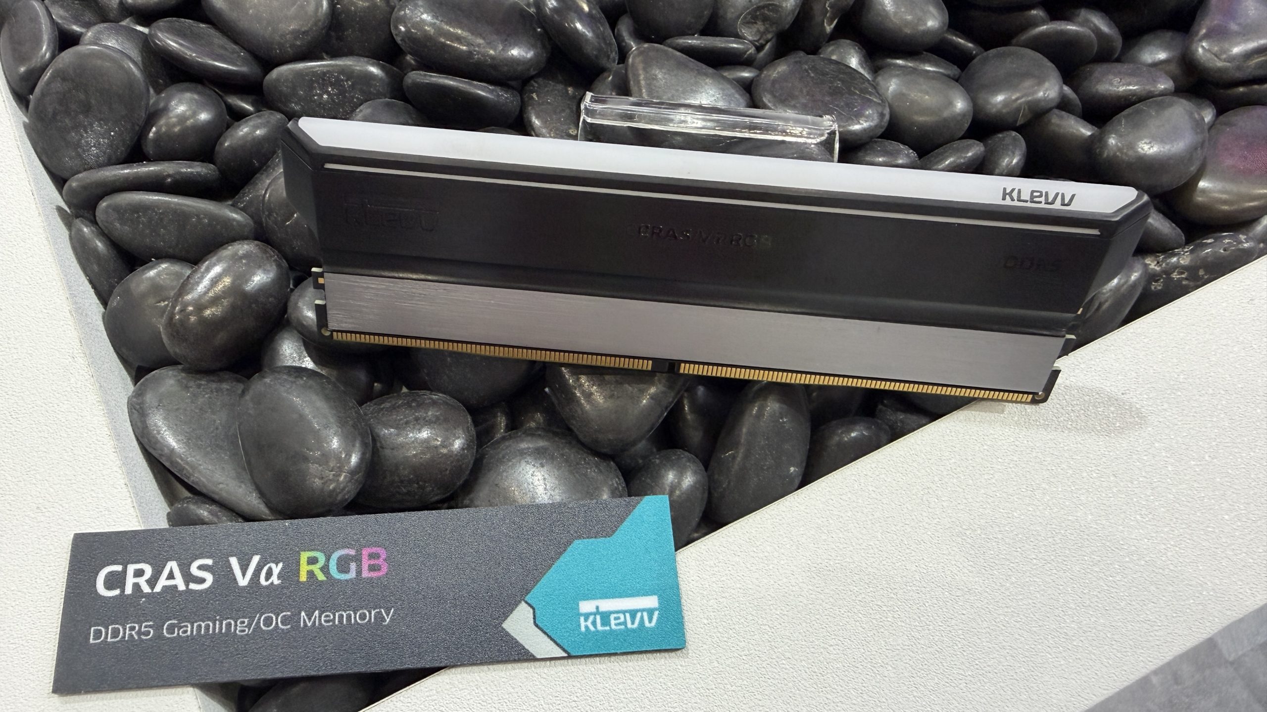 A KLEVV CRAS Vα RGB DDR5 Gaming/OC Memory module displayed on a bed of black stones with a label card.