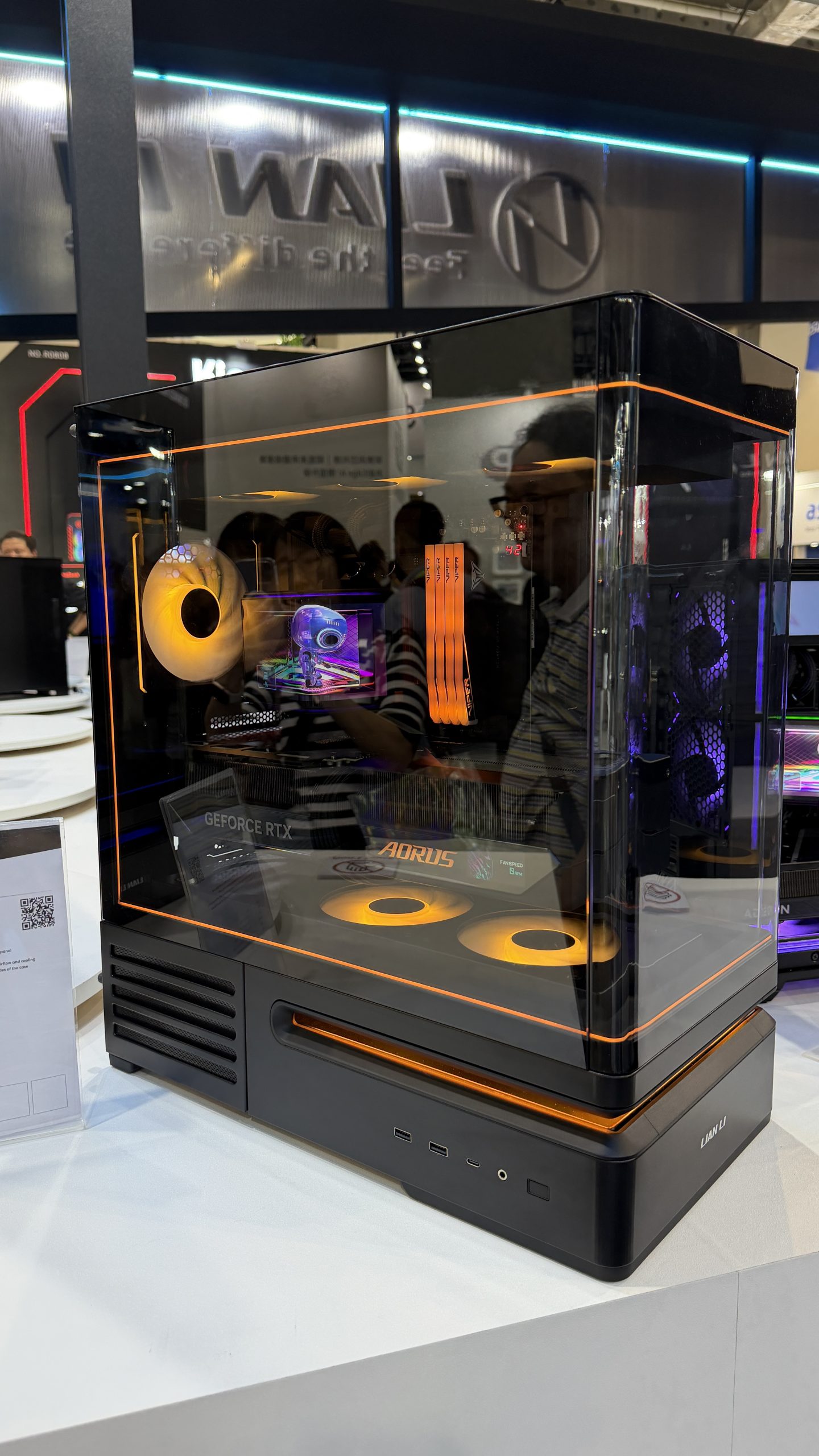 A custom PC build featuring a Lian Li case with orange LED lighting, visible AORUS components, and a 'GeForce RTX' graphics card on display at a tech event.