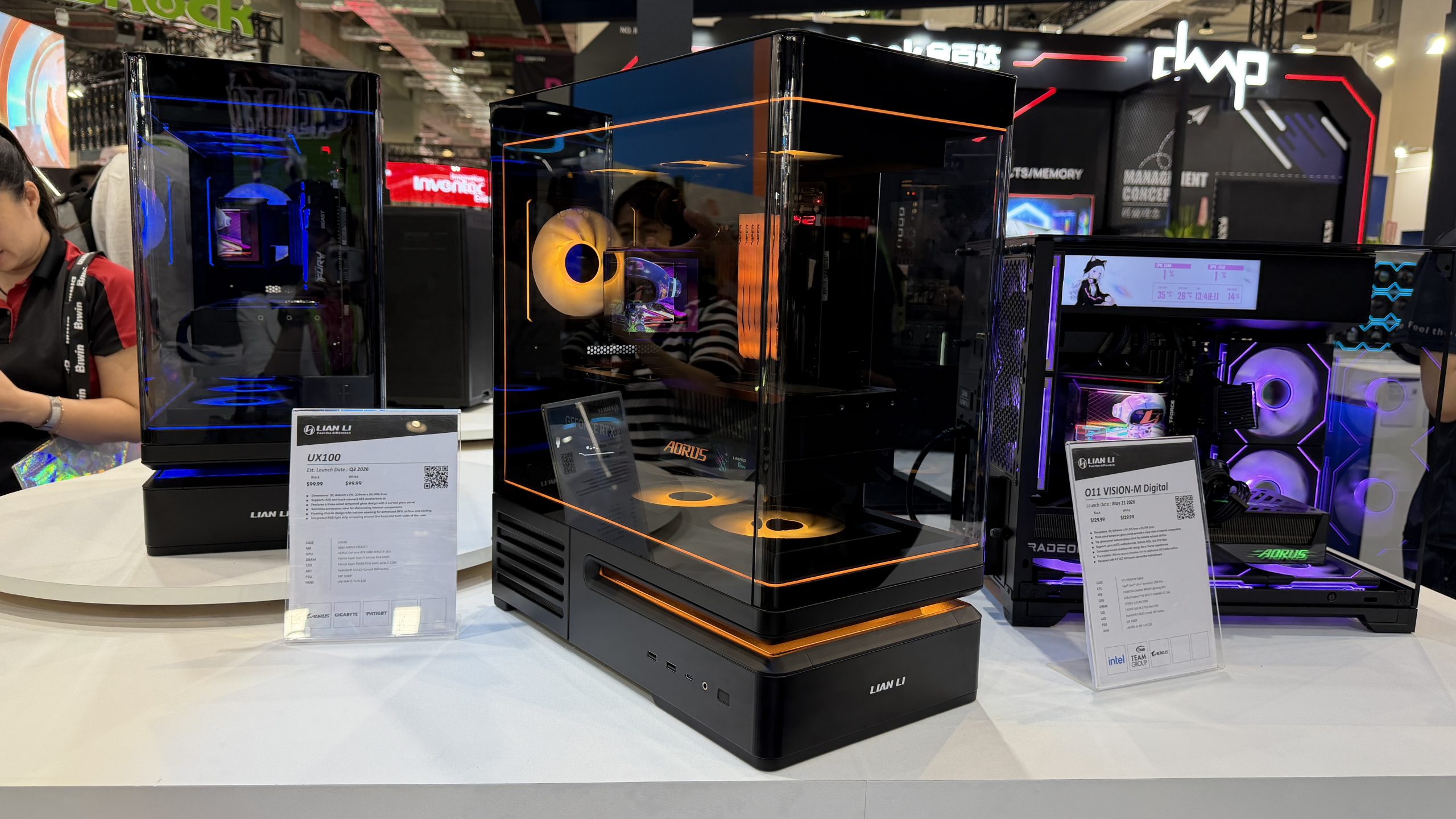 A display of Lian Li cases, including the 'UX100' and '011 VISION-M Digital,' featuring vibrant RGB lighting and visible AORUS branding, showcased in an exhibition setting.
