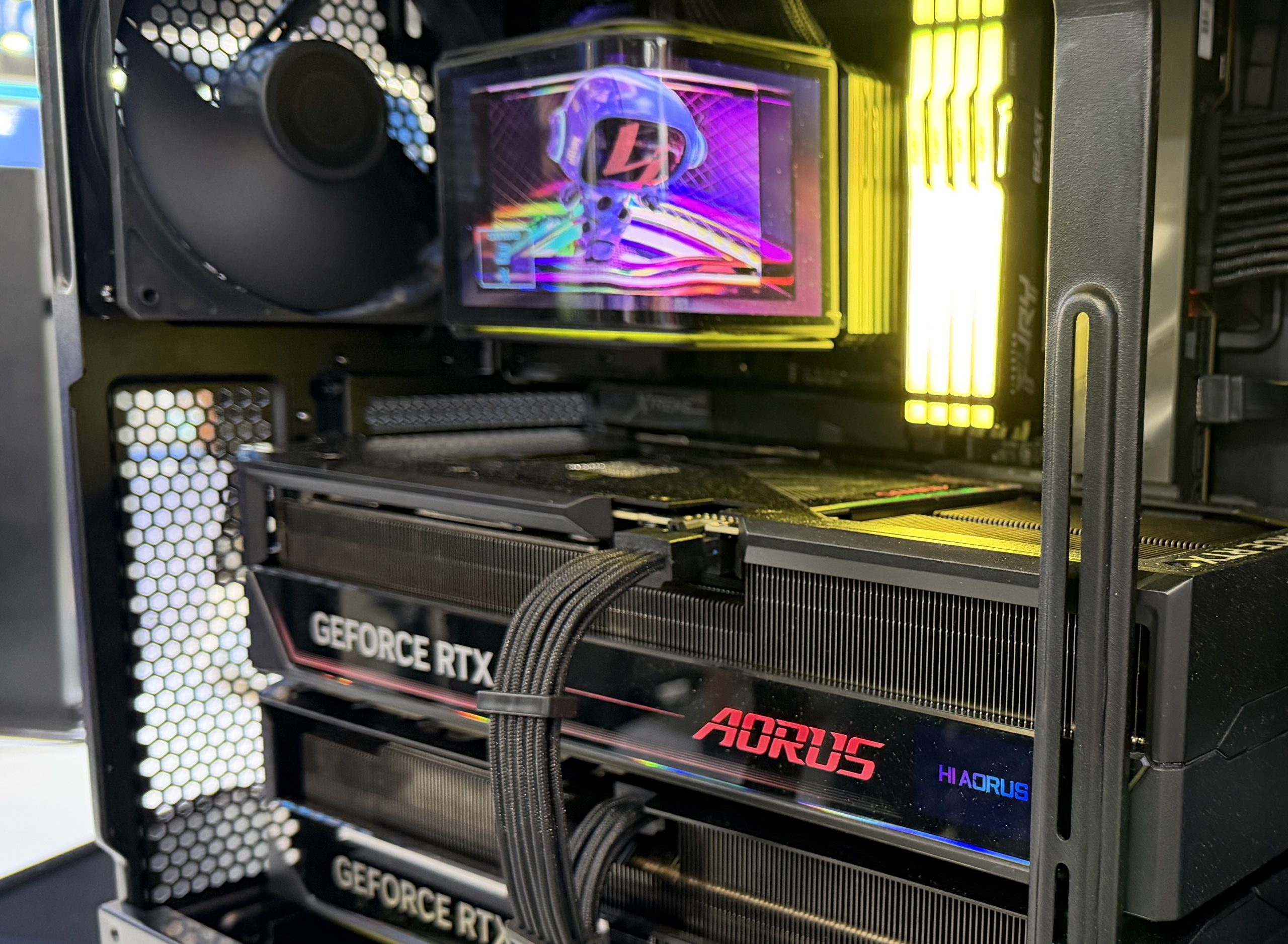An open PC case showcasing dual GeForce RTX graphics cards with AORUS branding, a colorful display on an AORUS water cooler, and illuminated RAM sticks.