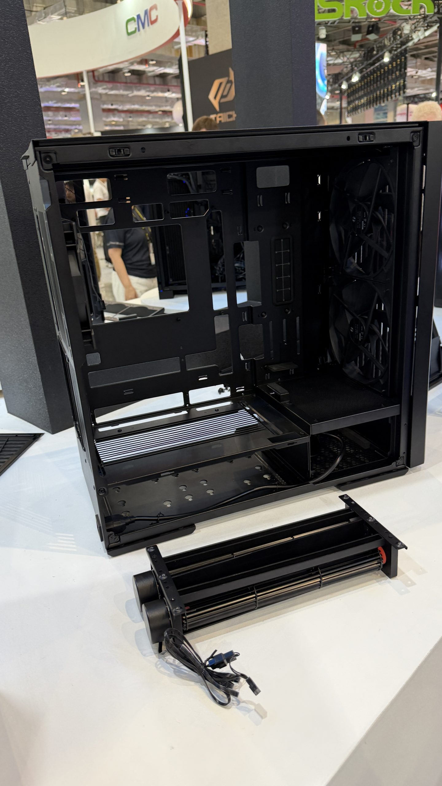 A black, open mid-tower PC case from Phanteks is displayed on a table, with a dual-fan radiator unit placed in front of it at a tech convention.