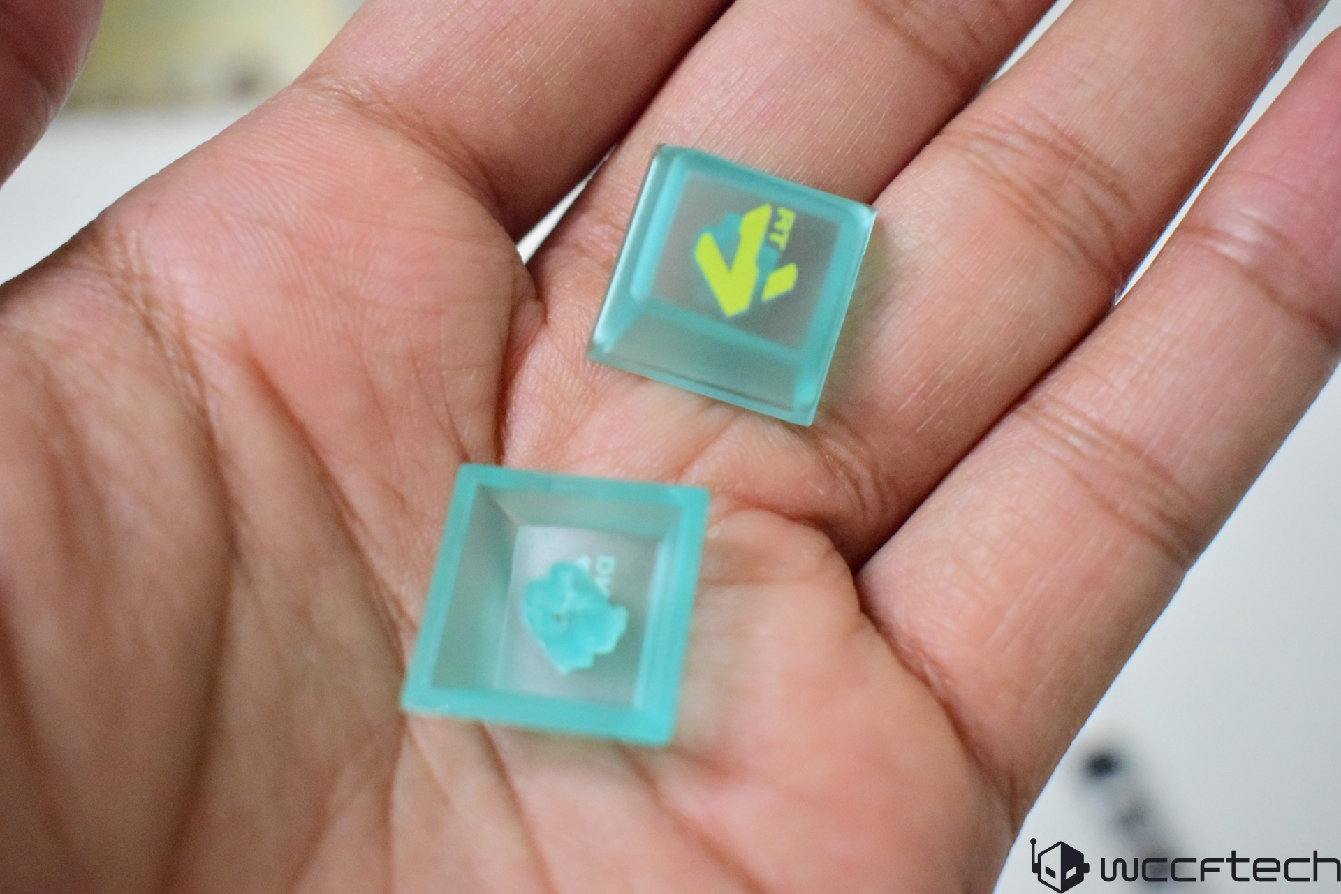 A hand holds two translucent teal keycaps, one featuring a yellow and green pattern with 'RT' on it.
