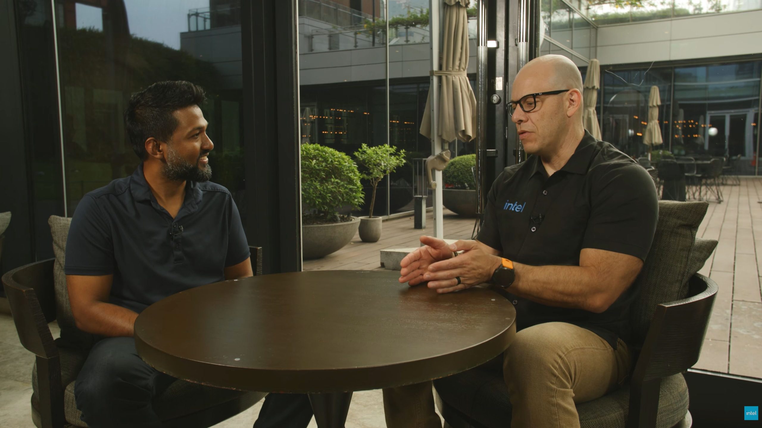 Two people sitting at a table having a discussion, with one wearing a polo shirt featuring the 'Intel' logo.