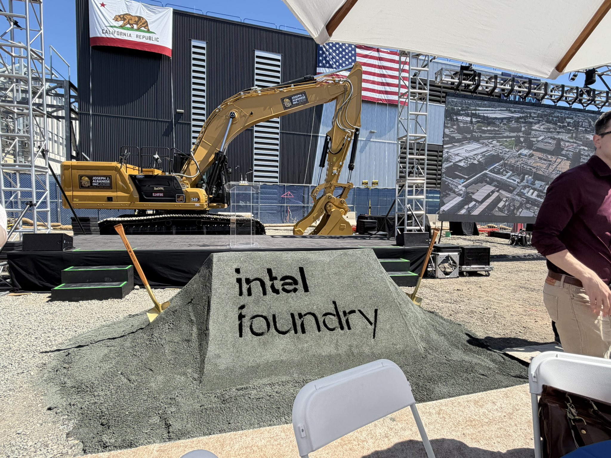A construction event with a Caterpillar 349 excavator and a mound of dirt displaying 'intel foundry' text, set against flags of California and the United States, with a screen showing an aerial view of a facility.