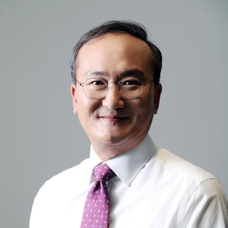 A man in glasses with a white shirt and pink tie smiling against a gray background.