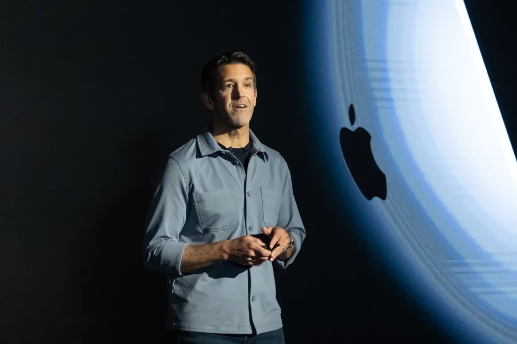 A presenter stands on stage beside a large Apple logo displayed on the screen.