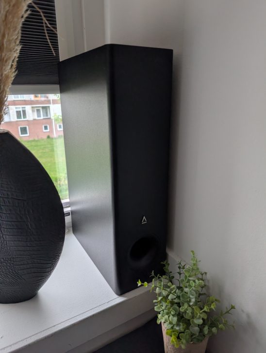 A black triangular speaker with a stylized logo is positioned on a windowsill next to a vase and a small potted plant.