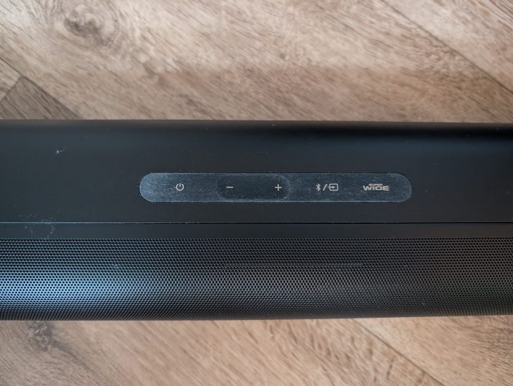A black soundbar with buttons labeled 'power,' 'minus,' 'plus,' 'Bluetooth,' and 'SUPER WIDE' is shown on a wooden surface.