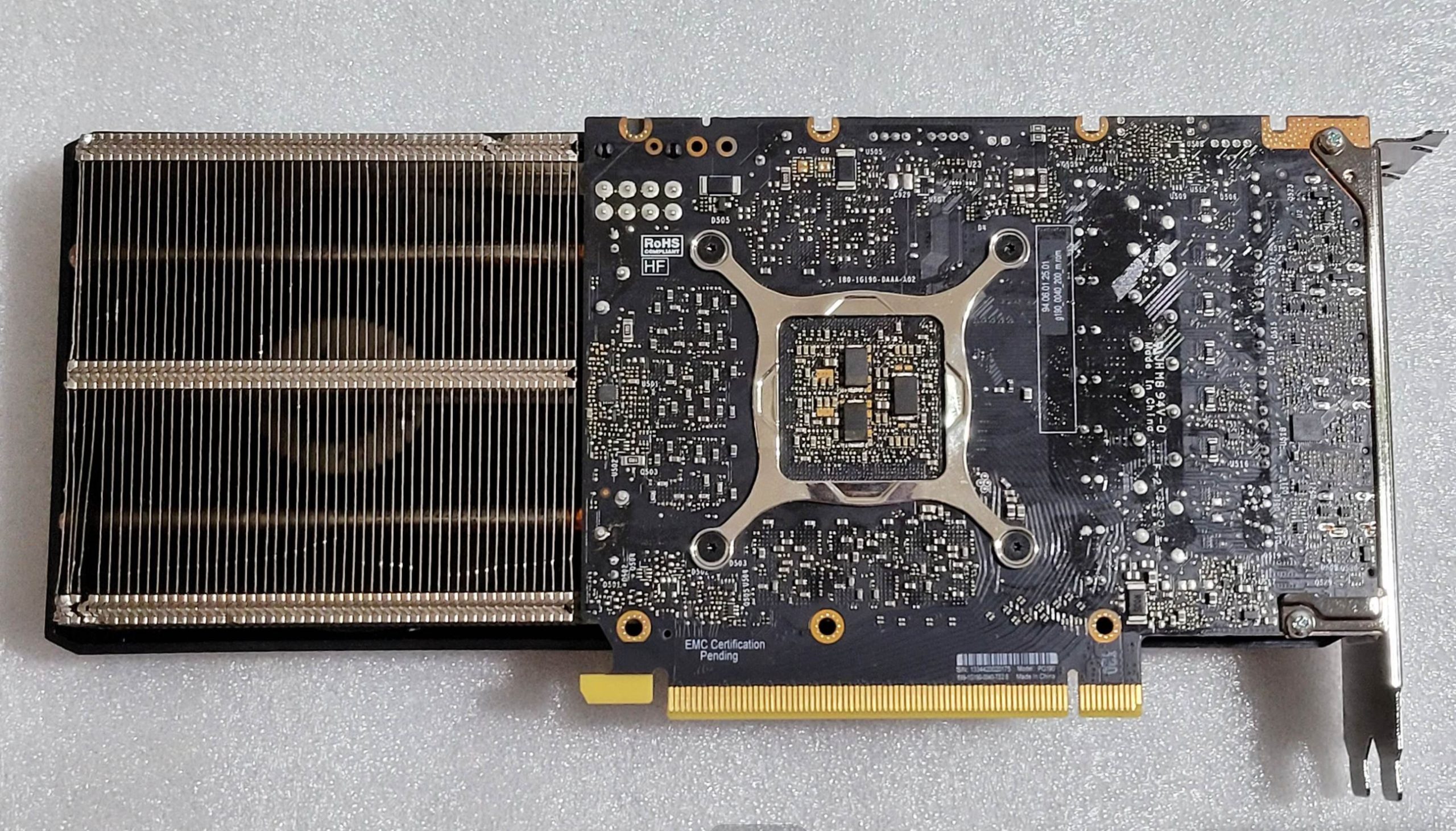 A back view of an unbranded graphics card showing the exposed circuit board and cooling fins, with 'EMC Certification Pending' text visible near the PCI-Express connector.