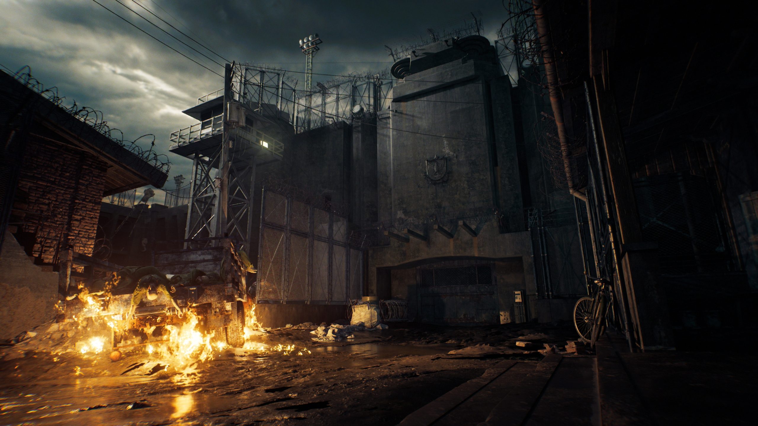 A scene from a game showing a burning truck in front of a fortified, barbed-wire prison wall under a cloudy sky.