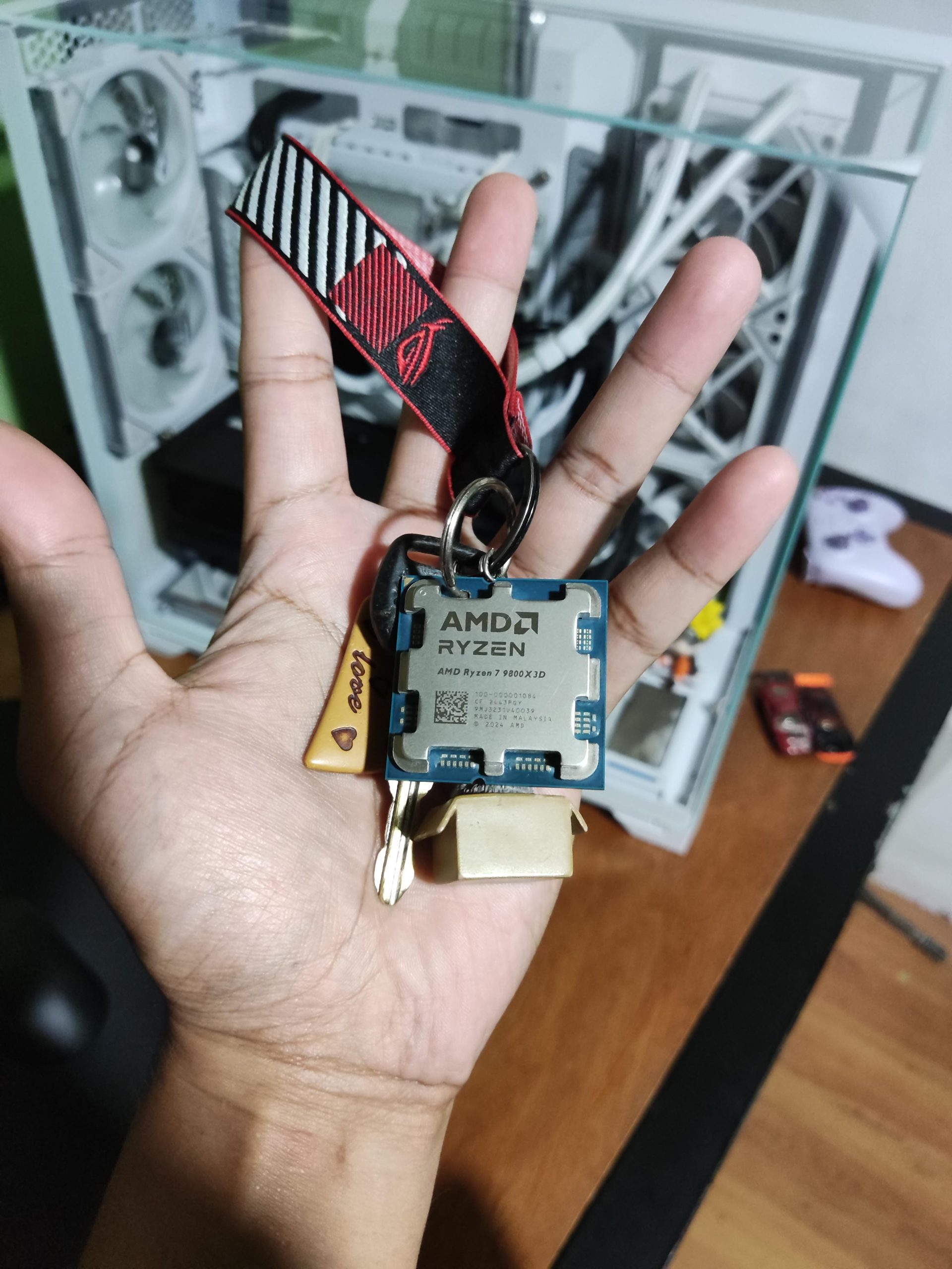 A person holding an AMD Ryzen 7 7800X3D processor keychain with a gaming PC in the background.