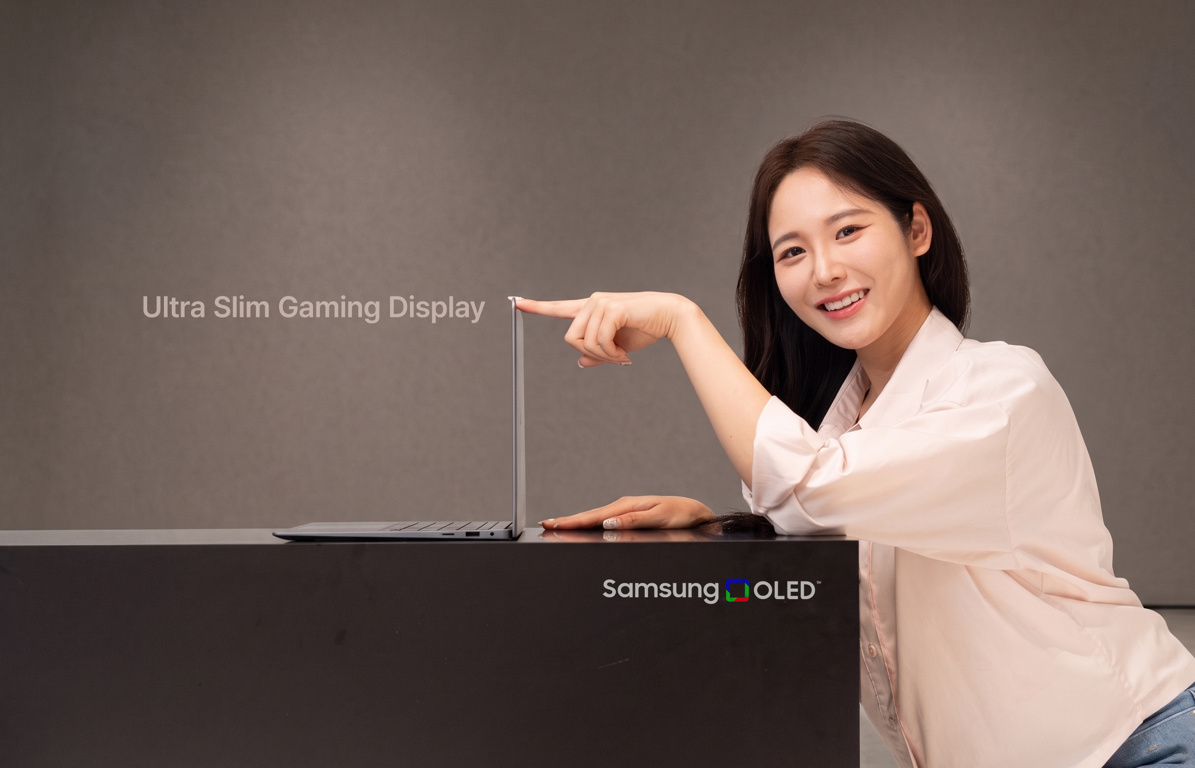 A person smiles and points at an ultra-thin laptop screen next to the text 'Ultra Slim Gaming Display,' with 'Samsung OLED' visible on the table.