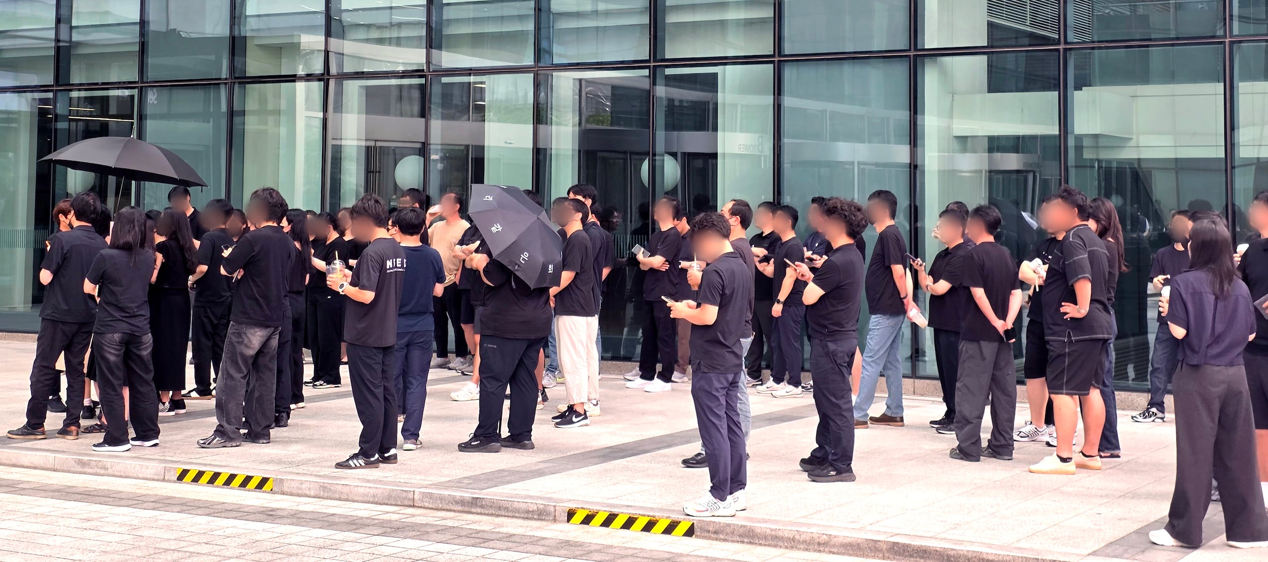 A group of people in black clothing gather outside a glass-fronted building, with some holding umbrellas and drinks.